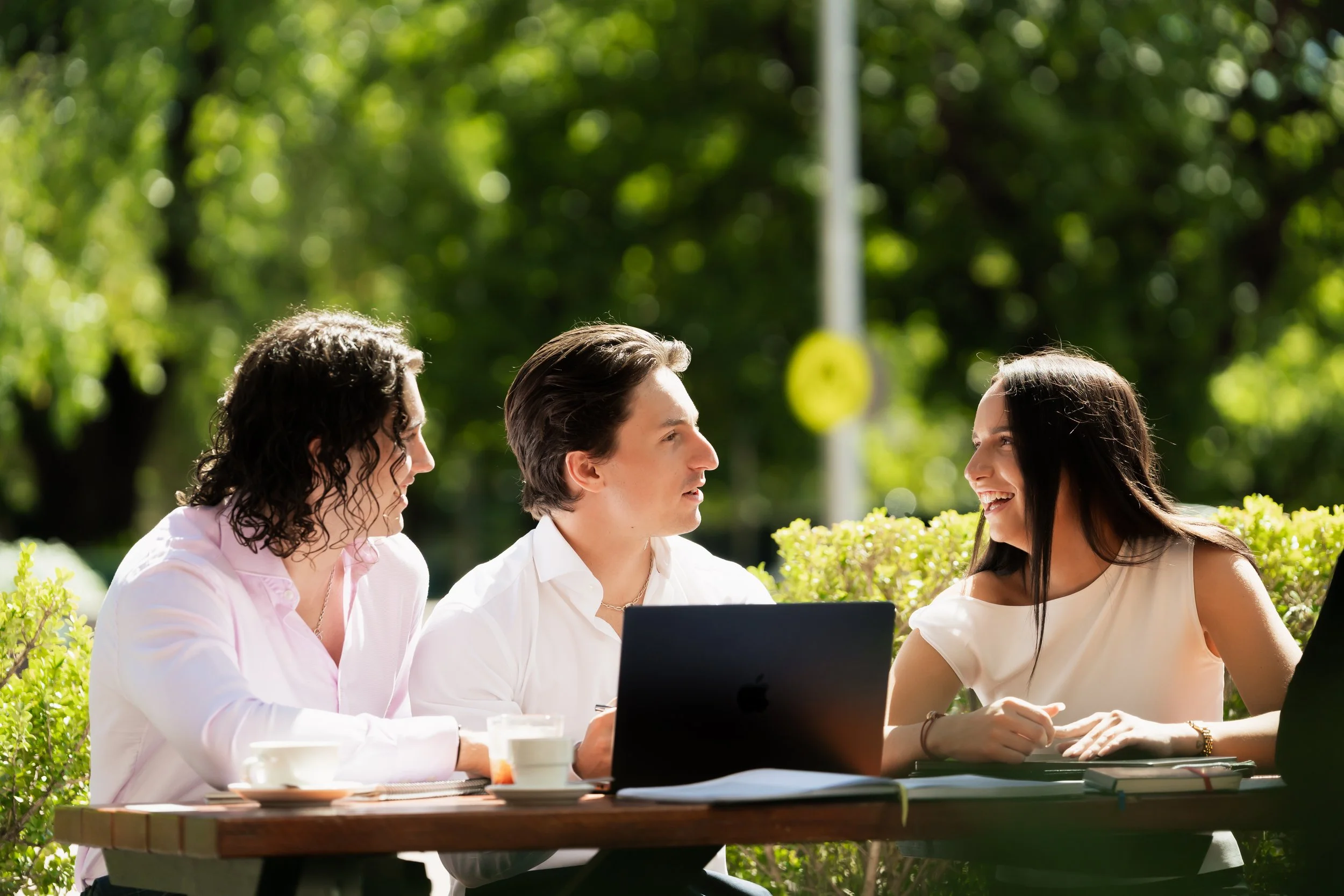 Four professional adults sitting at an outdoor table, engaged in conversation on a sunny day surrounded by greenery having a business meeting and planning financial services 