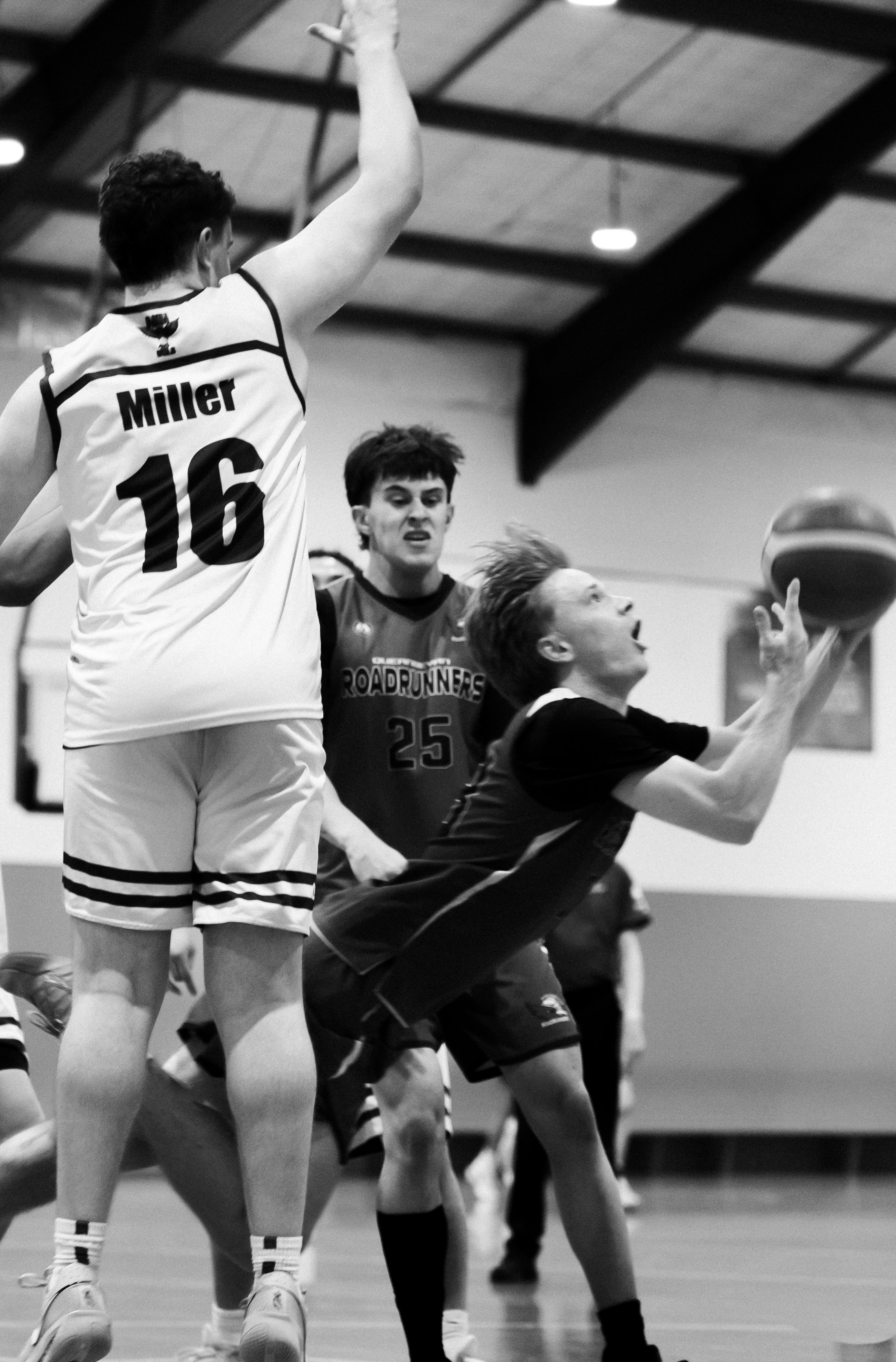 A black and white photo of boys playing basketball in a gymnasium. One player is jumping and holding the basketball, while another is defending and reaching for the ball. ANU australian national university mens basketball team sports photography