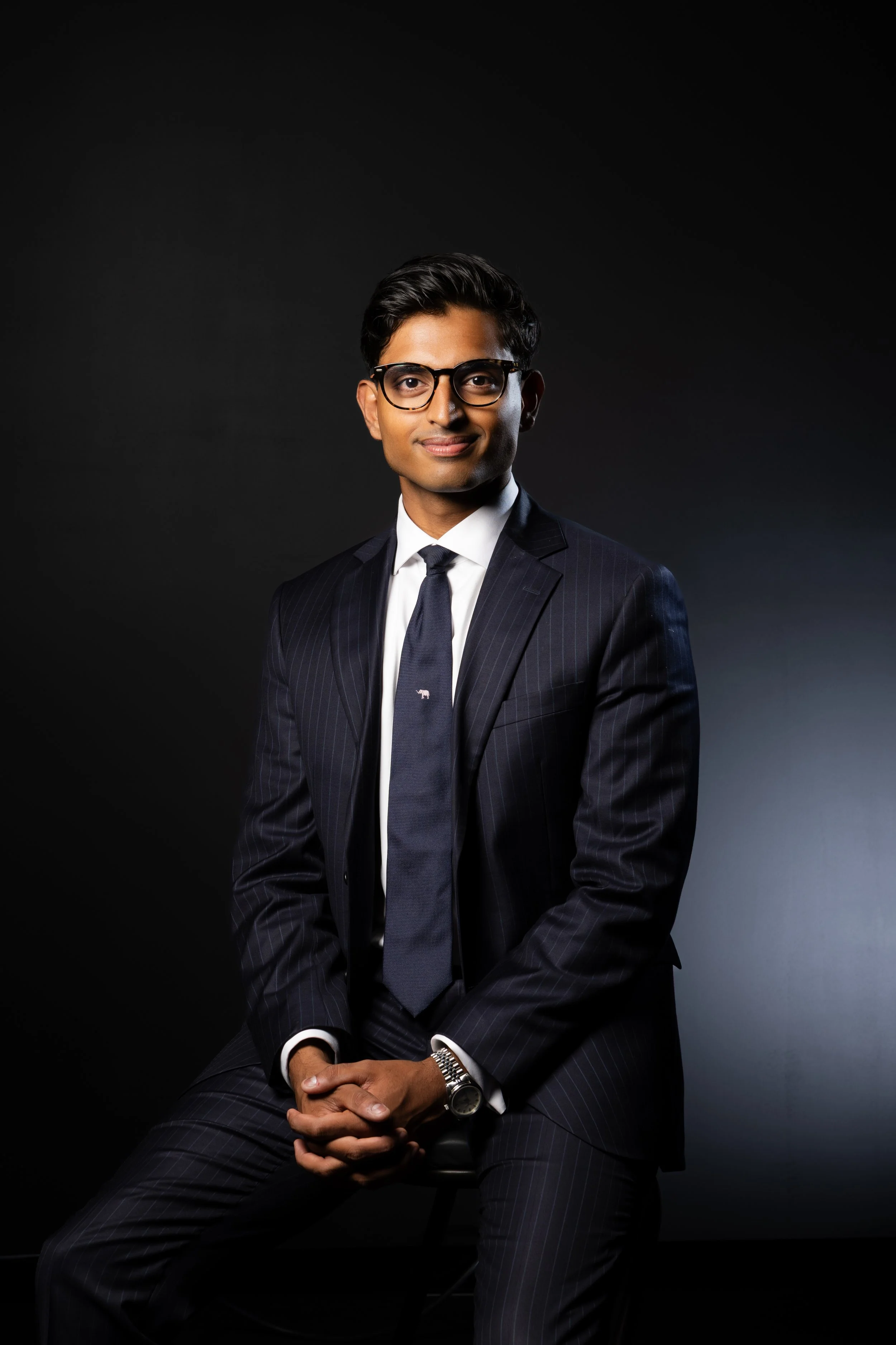 A young man in a dark pinstripe suit, white shirt, and dark tie sitting against a dark background. He is wearing glasses and has a watch on his left wrist, with his hands clasped. Real estate sales agent headshot and studio portrait