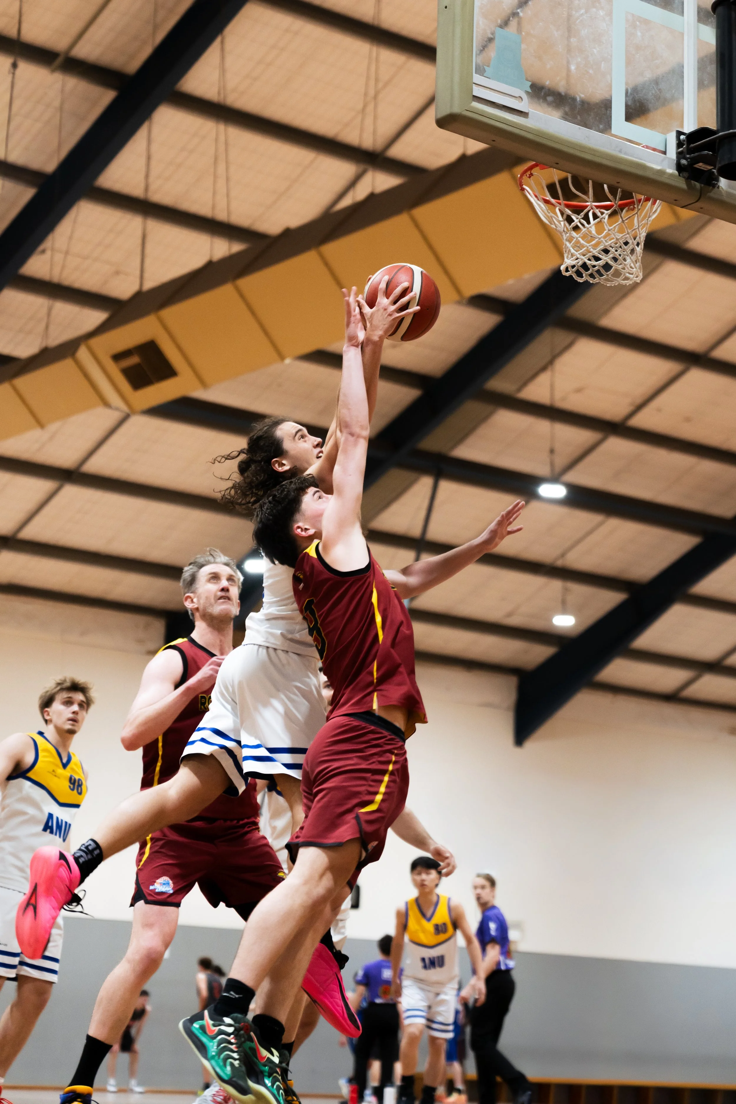 basketball players in mid-air competing for the ball near the hoop during a game in an indoor gymnasium. ANU australian national university basketball mens team sports photography