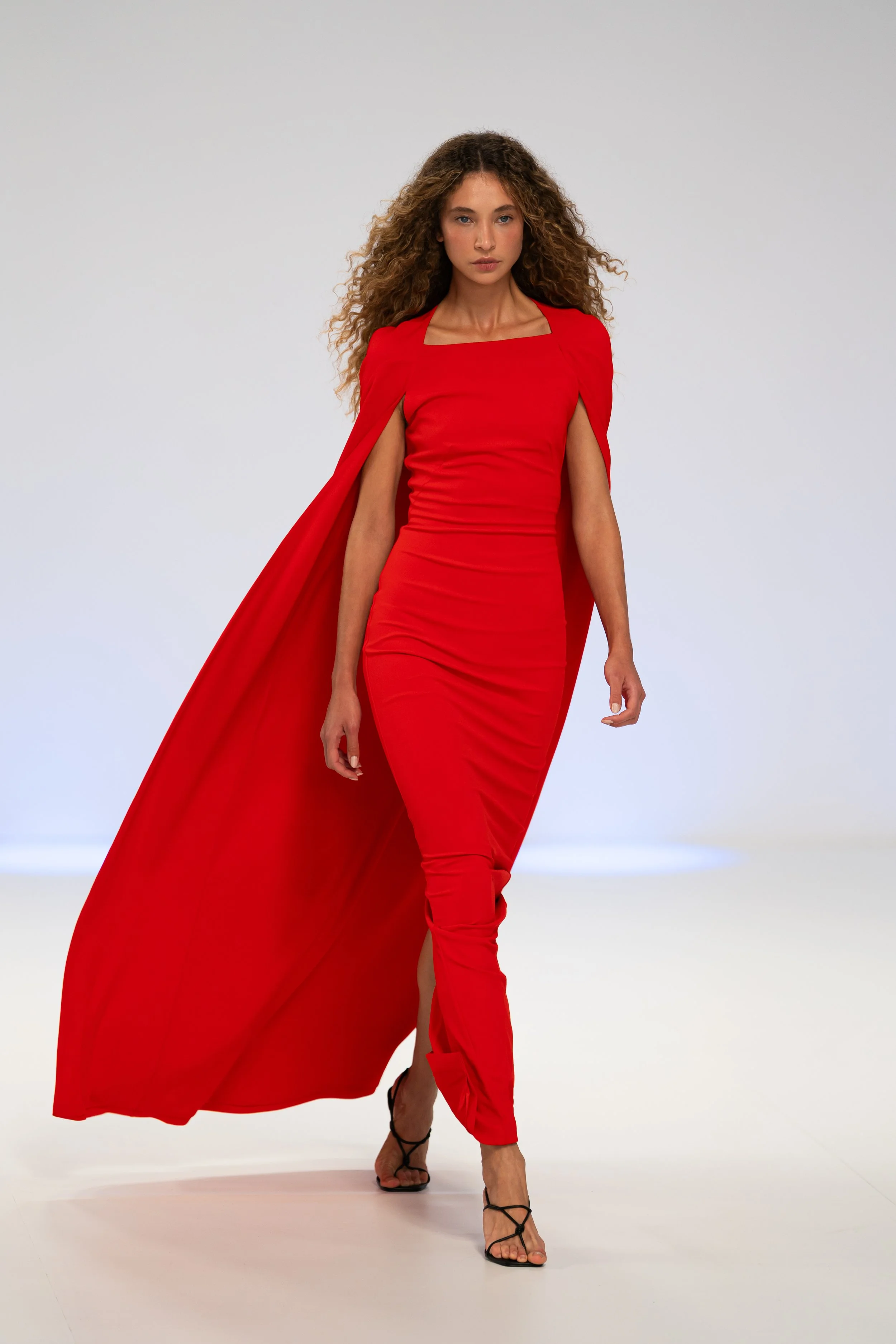 A woman walking on a runway in a long red gown with a cape, wearing strappy black high heels, with a neutral gray background. australian fashion week 2026 effie kats runway show the edit runway show