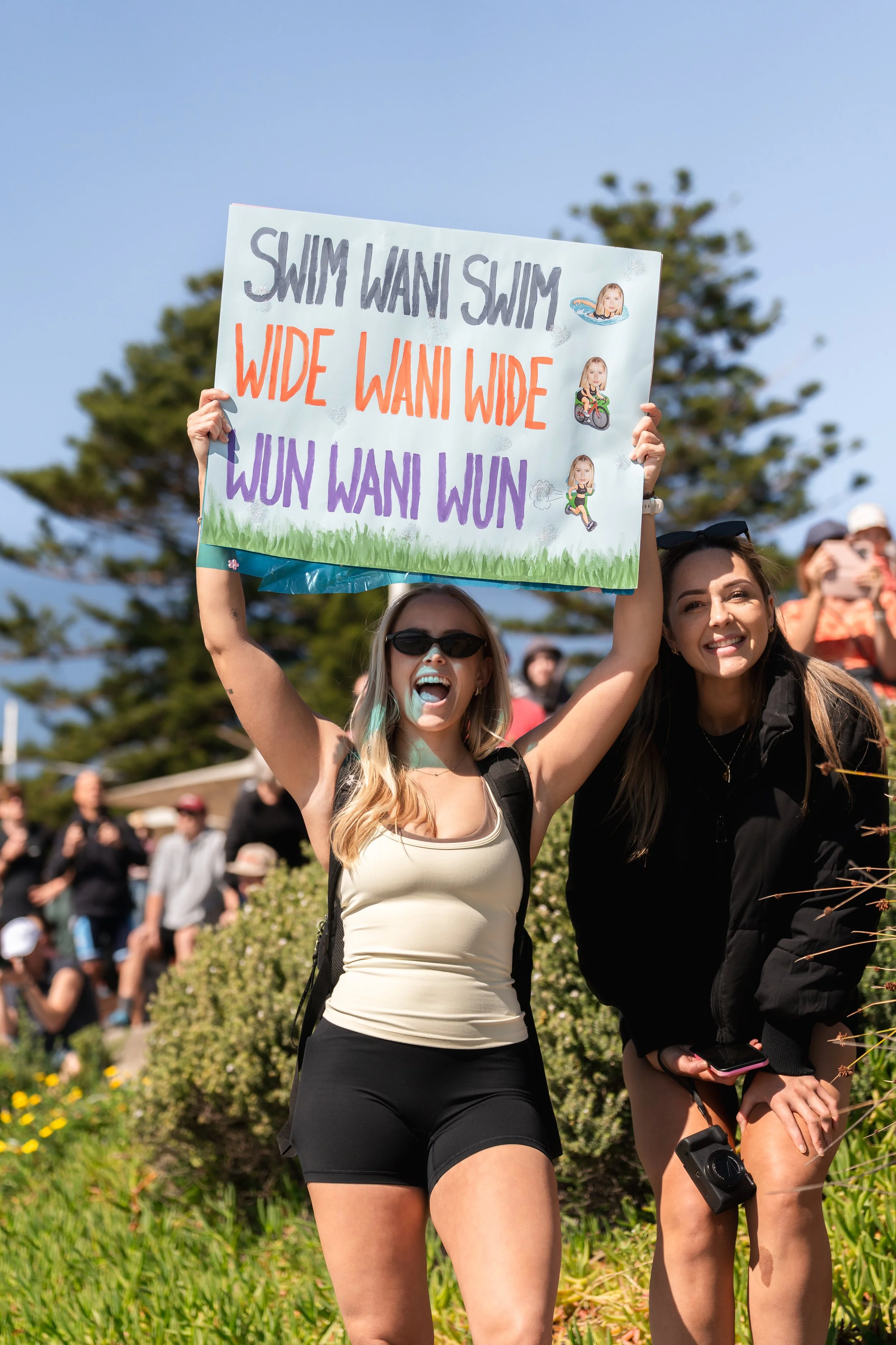 Two women are holding a colorful sign with text about swimming, walking, and running, during a daytime outdoor event with many people in the background. wollongong triathlon 2026 sports photography