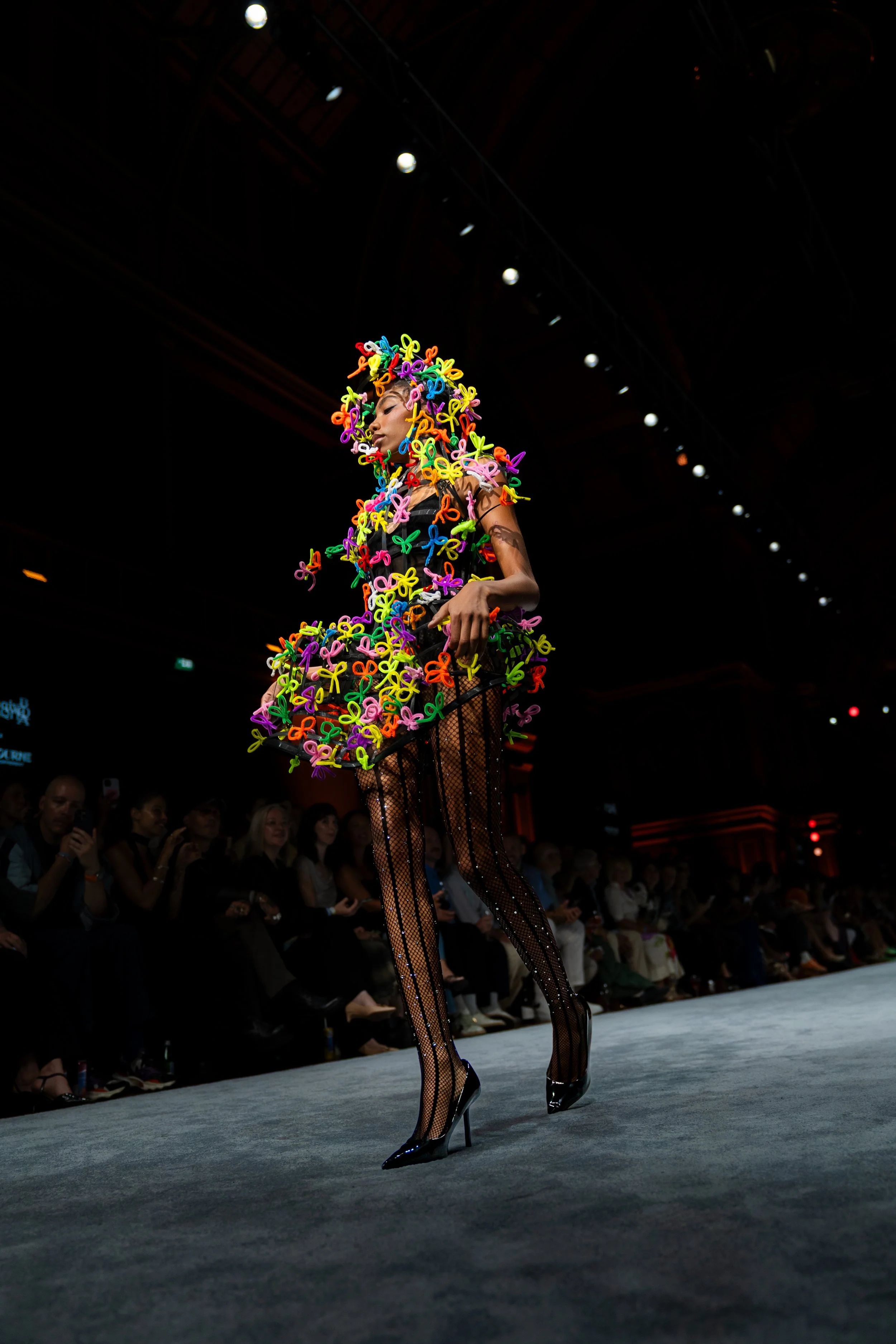 A fashion model walking down the runway during a fashion show, wearing a colorful outfit adorned with multicolored bows and black fishnet stockings. The audience watches from the background. melbourne fashion festival Baaqiy runway show
