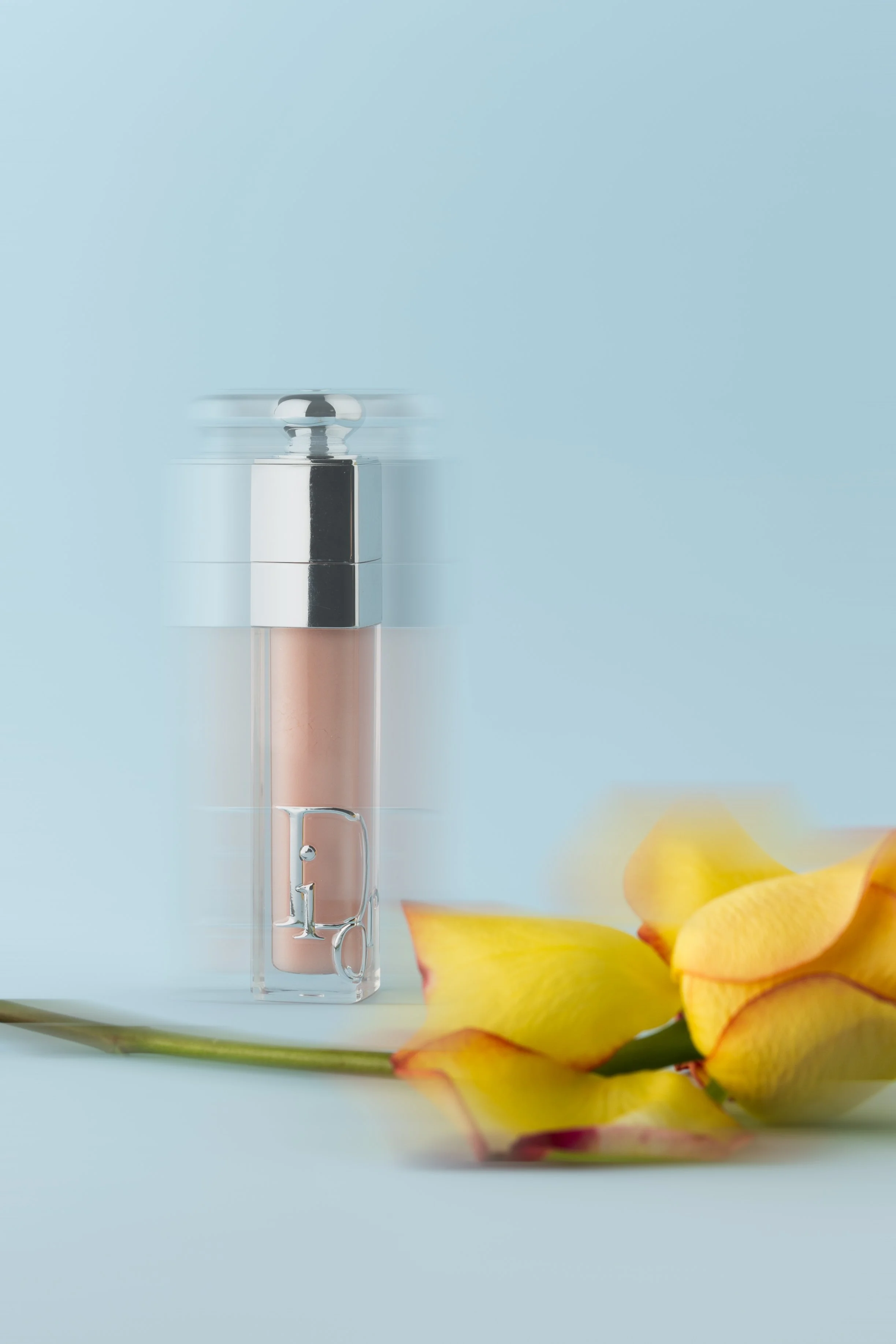 A bottle of perfume with a metallic cap, placed on a small stand, and yellow rose petals on a yellow plate against a light blue background. dior lip gloss product photography