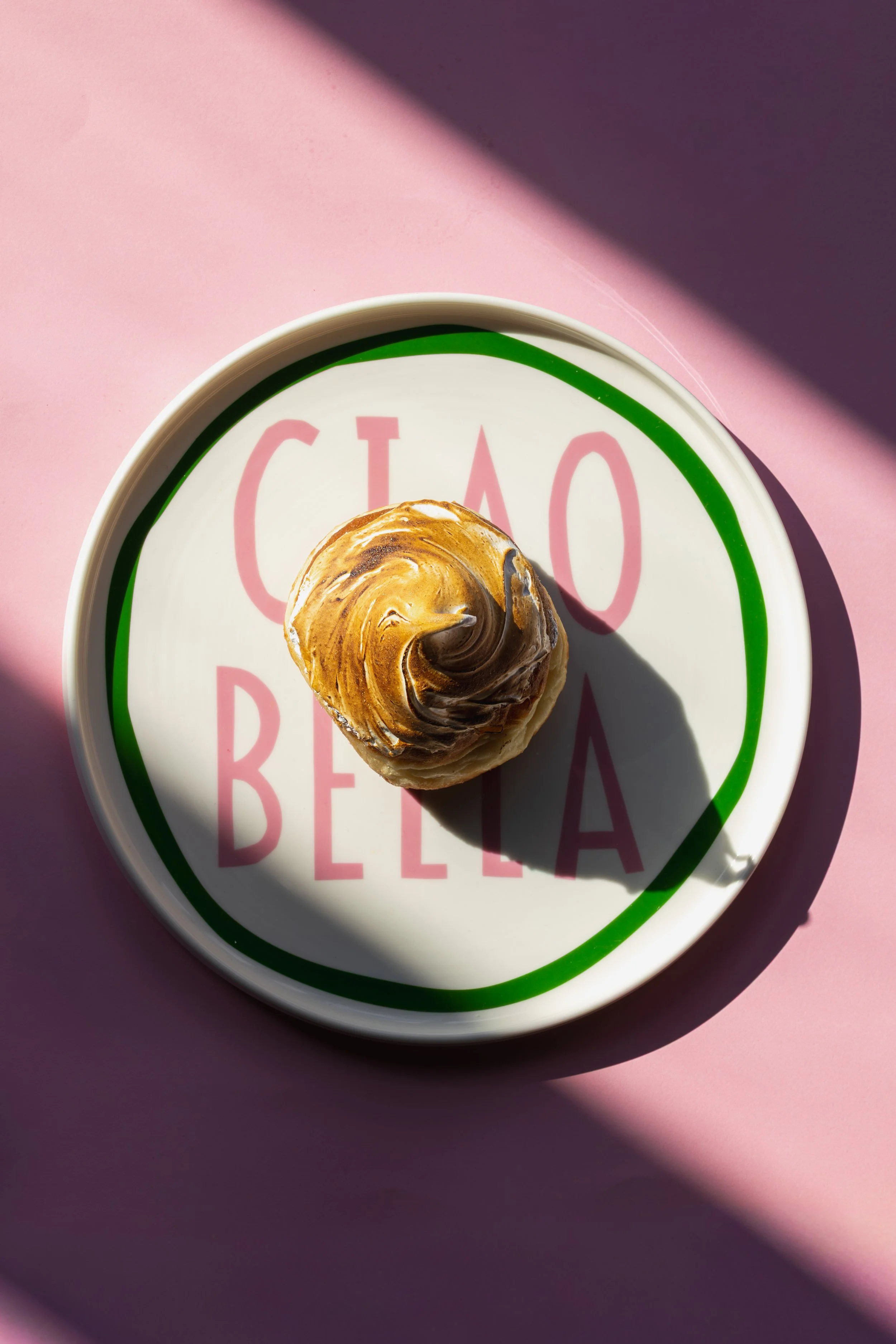 A close-up of a cupcake with swirled frosting in a swirl pattern on a plate with pink and green text, set on a pink background with shadows. round of house ciao bella plate product photography