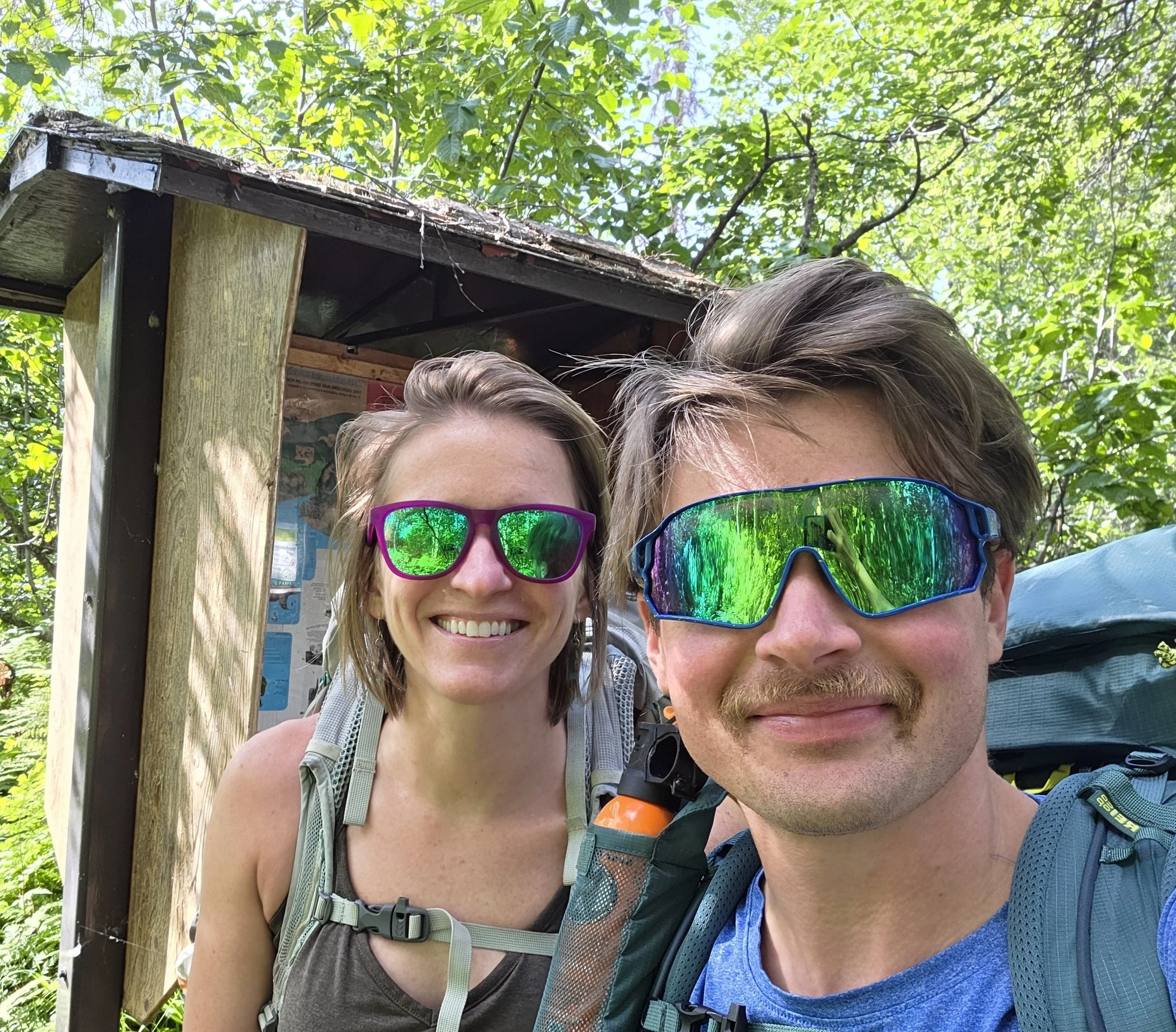 Close-up of two people taking a selfie outdoors with green trees in the background. They are wearing sunglasses and hiking gear.