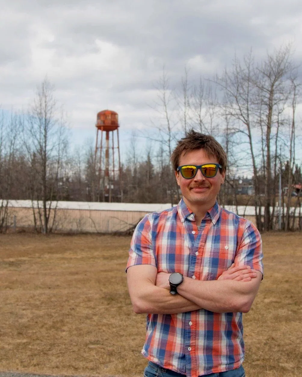 A man with sunglasses and a plaid shirt standing outdoors with arms crossed, a water tower, leafless trees, and a cloudy sky in the background.