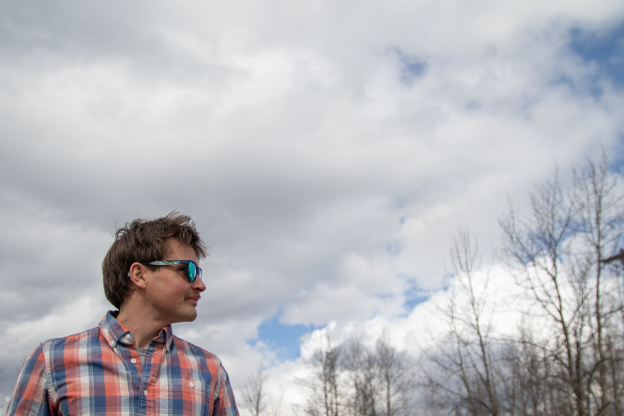 Side profile of a young man with brown hair and wearing reflective sunglasses and a plaid shirt, standing outdoors under a cloudy sky with leafless trees in the background.