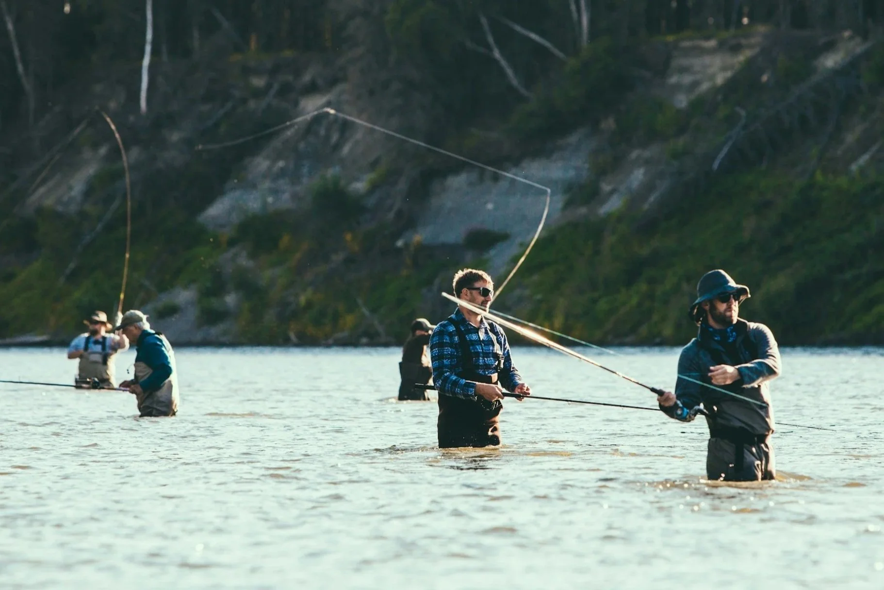 Multiple people fishing in a body of water, standing in waist-high water, with a scenic background of a forested hill.