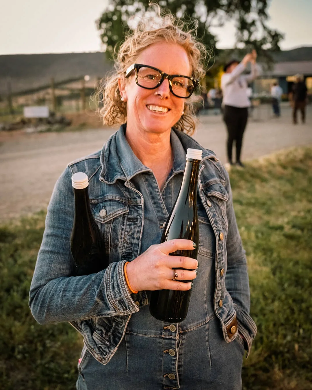 A smiling woman with curly blonde hair, black glasses, and a denim jacket holding two dark bottles outdoors during sunset.