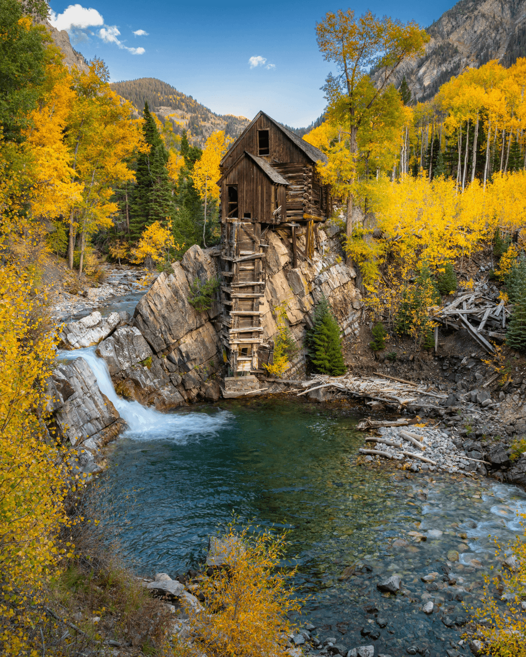 An old wooden cabin built on a rocky cliff over a river, surrounded by autumn-colored trees and mountains in the background.