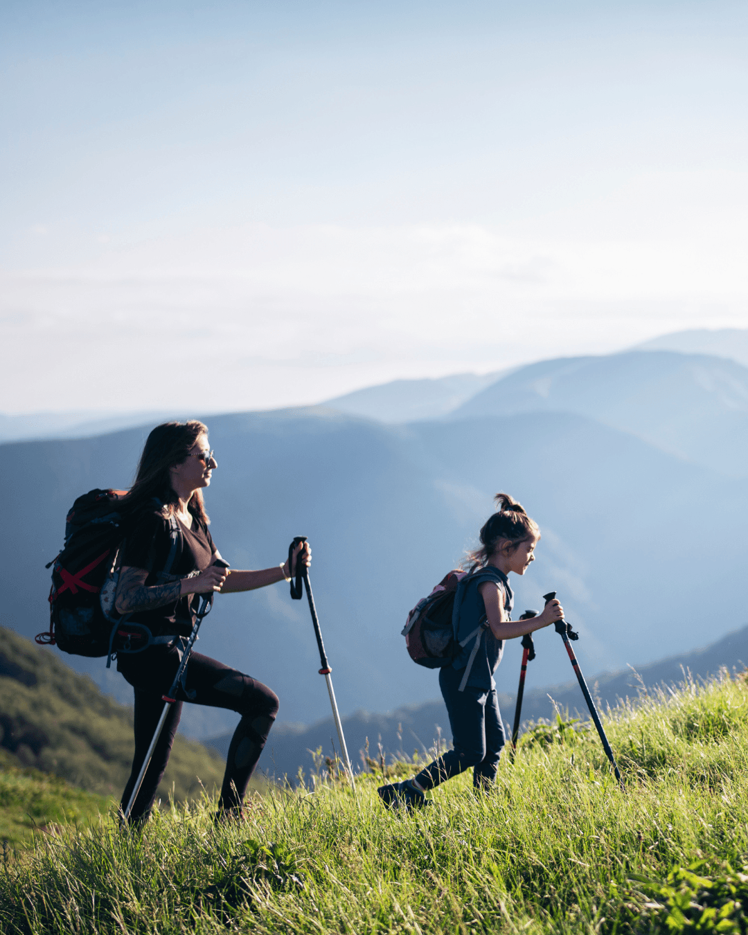 A woman and a girl hiking on a grassy mountain trail with a scenic mountain range in the background.