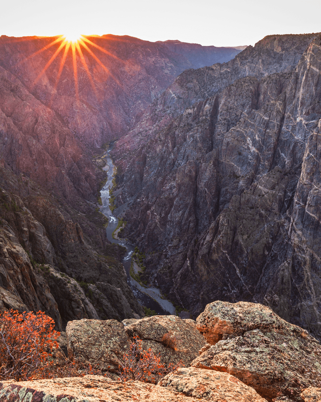 Sunset over rugged canyon with river flowing through it, viewed from rocky cliff edge.