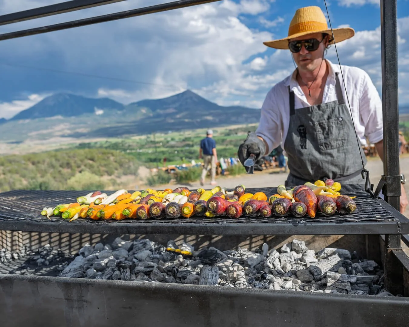 Person wearing a wide-brimmed hat, sunglasses, and apron grilling colorful vegetables on a barbecue outdoors with mountains in the background.