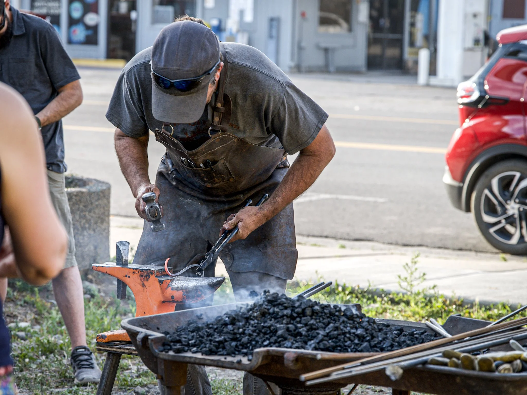 A man wearing a gray cap, sunglasses, and work apron is forging metal with a hammer and anvil outdoors near a parking lot.