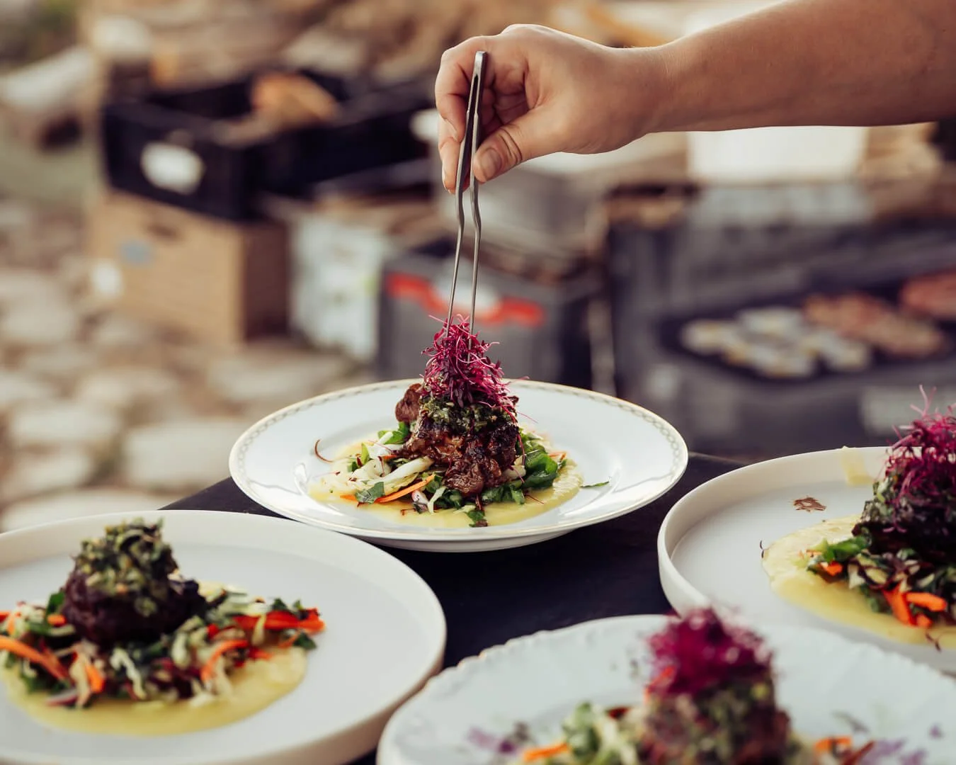 A person using tongs to place purple shredded garnish on a plated dish with meat and vegetables, with several similar dishes on a dark table.
