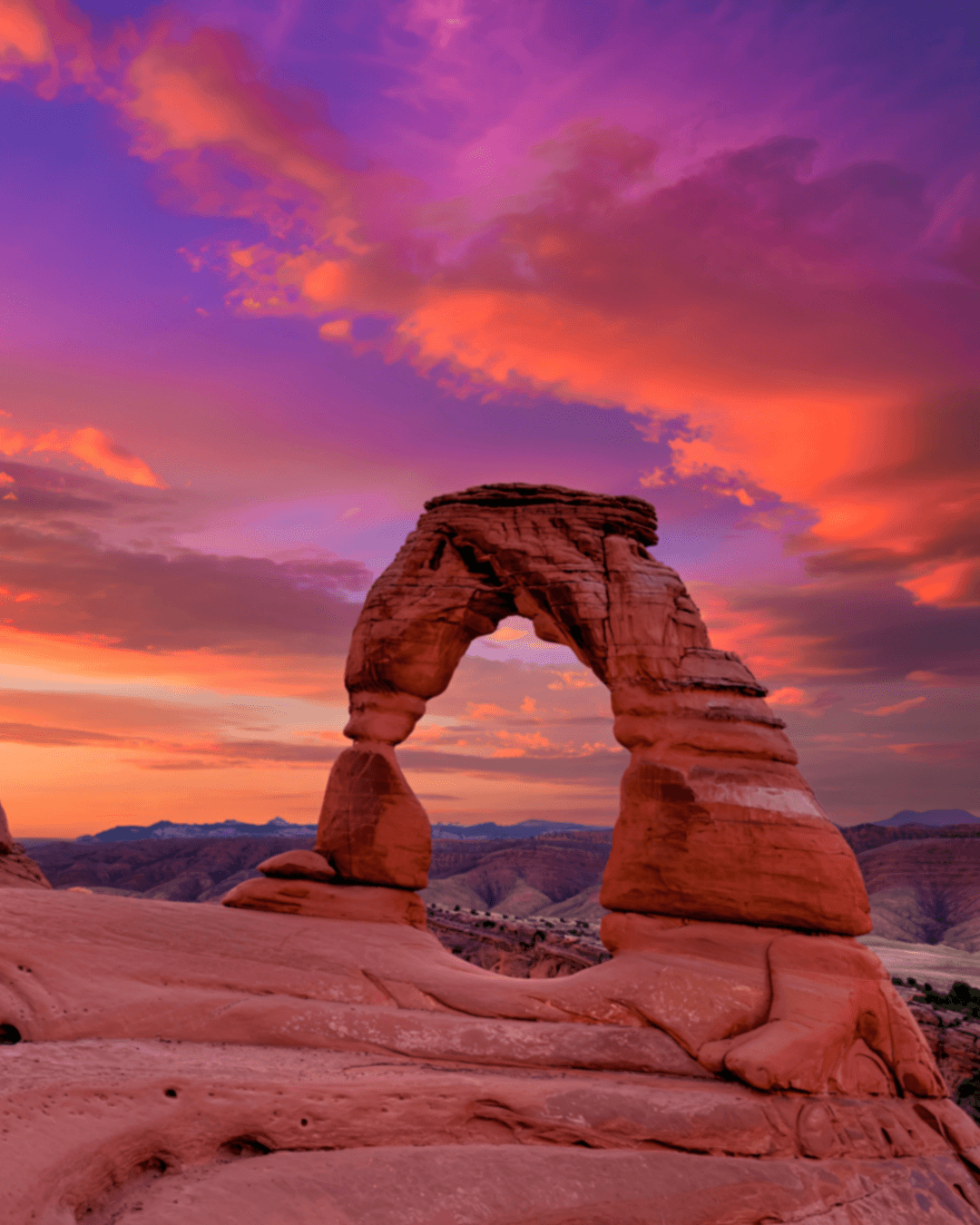 The Delicate Arch, a natural rock formation, during sunset with a colorful sky filled with pink, purple, and orange clouds.