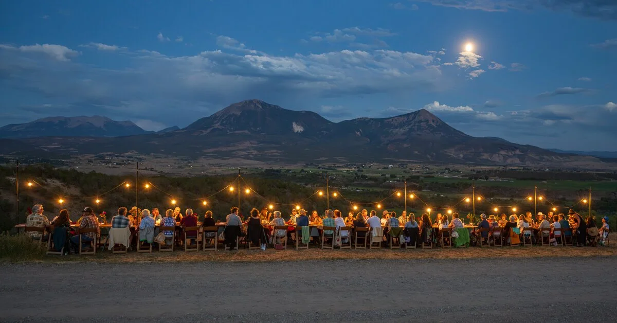 People seated at a long outdoor table under string lights during dusk, with mountains and a cloudy sky with a bright moon in the background.
