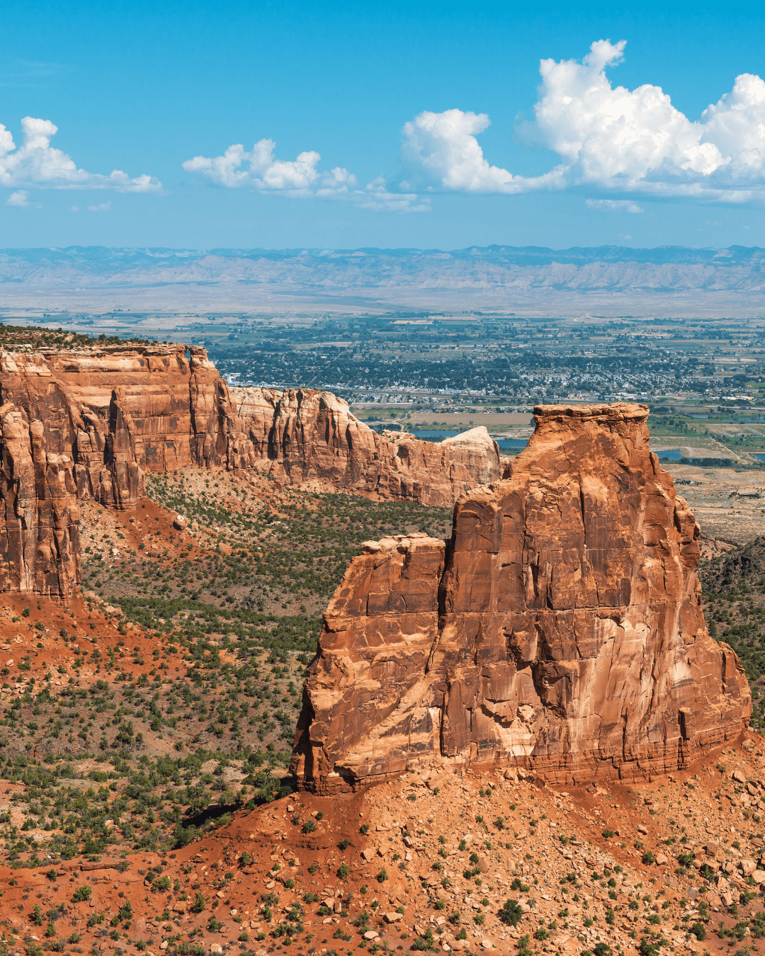 Red rock formations in a desert landscape with a city and mountains in the background under a partly cloudy sky.