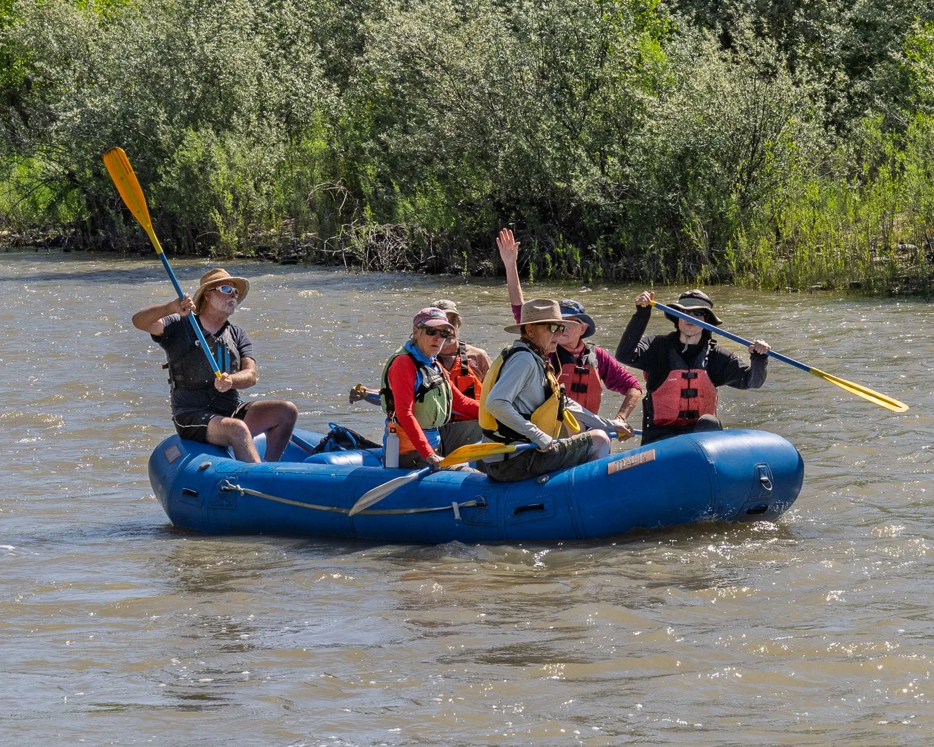 Group of people on a blue inflatable raft paddling on a river surrounded by green trees, wearing life jackets, hats, and sunglasses.