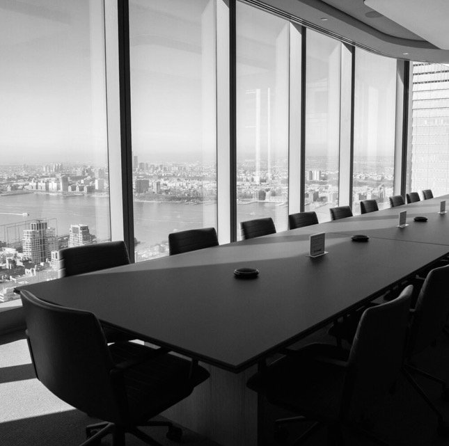 A modern conference room with a long table and office chairs, overlooking a cityscape through large windows.