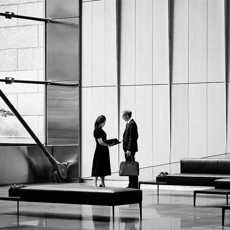 A woman and a man are standing in a modern office lobby. The woman is showing the man a folder or document. The man is holding a briefcase and looking at the woman. The lobby has a large wall with vertical panels, a bench, and a sofa.