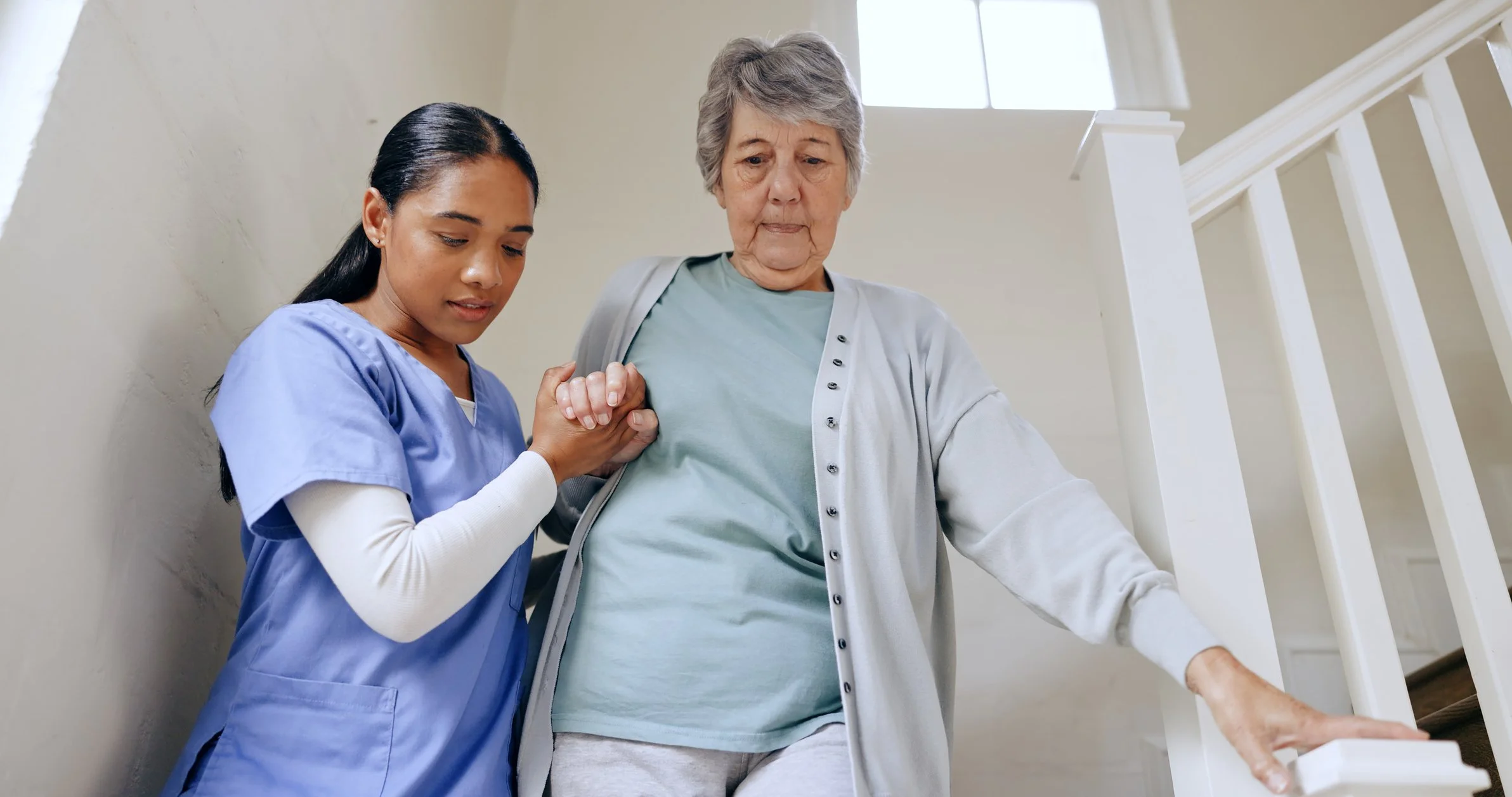 A young female healthcare worker helping an elderly woman as she walks down the stairs inside a home.