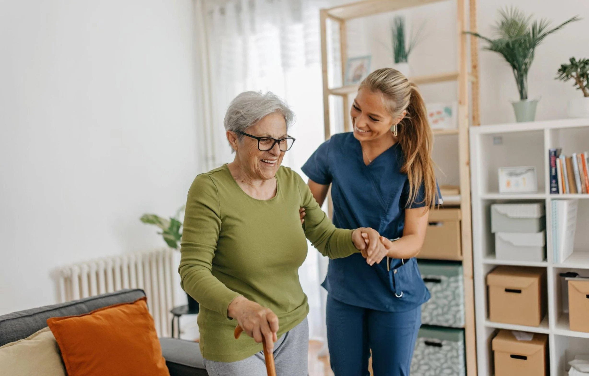 An elderly woman with gray hair and glasses, holding a cane and smiling, being assisted by a young woman in medical scrubs. They are in a bright living room with plants and bookshelves.