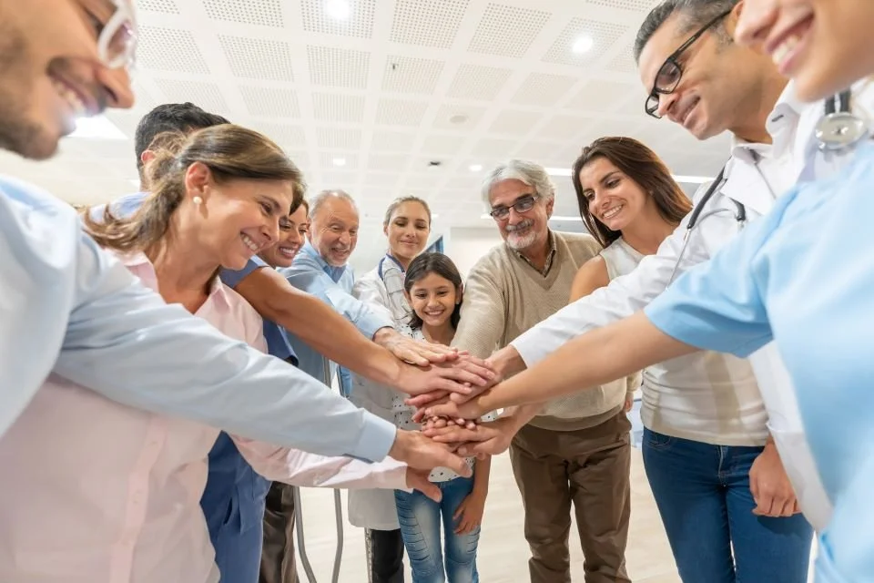 A diverse group of medical professionals and a child wearing scrubs and lab coats, placing their hands together in a gesture of teamwork and unity in a healthcare setting.