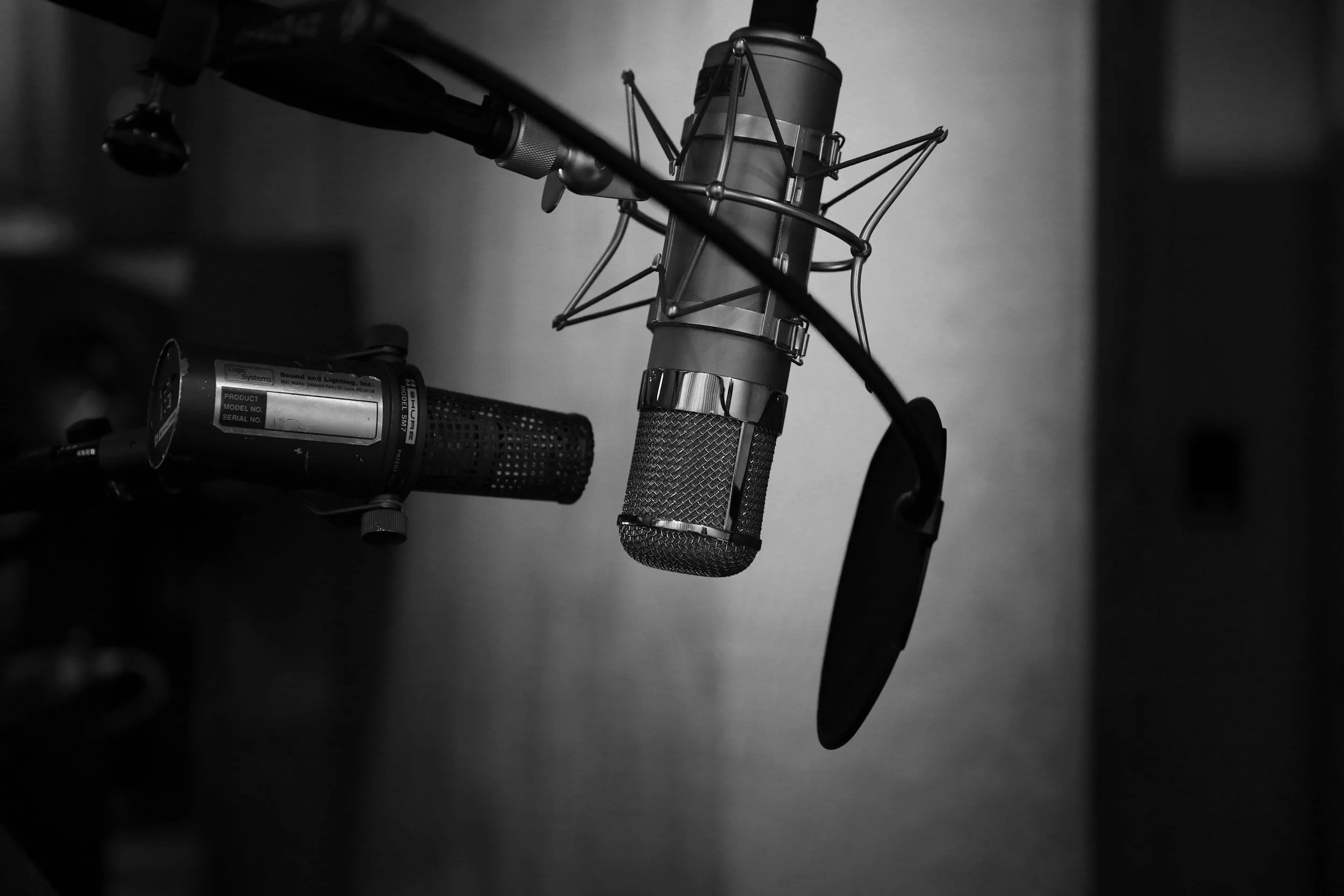 A black and white photo of a vintage studio microphone with a shock mount, with a flash of light reflecting off its metal parts, mounted on a stand, part of a professional recording setup.