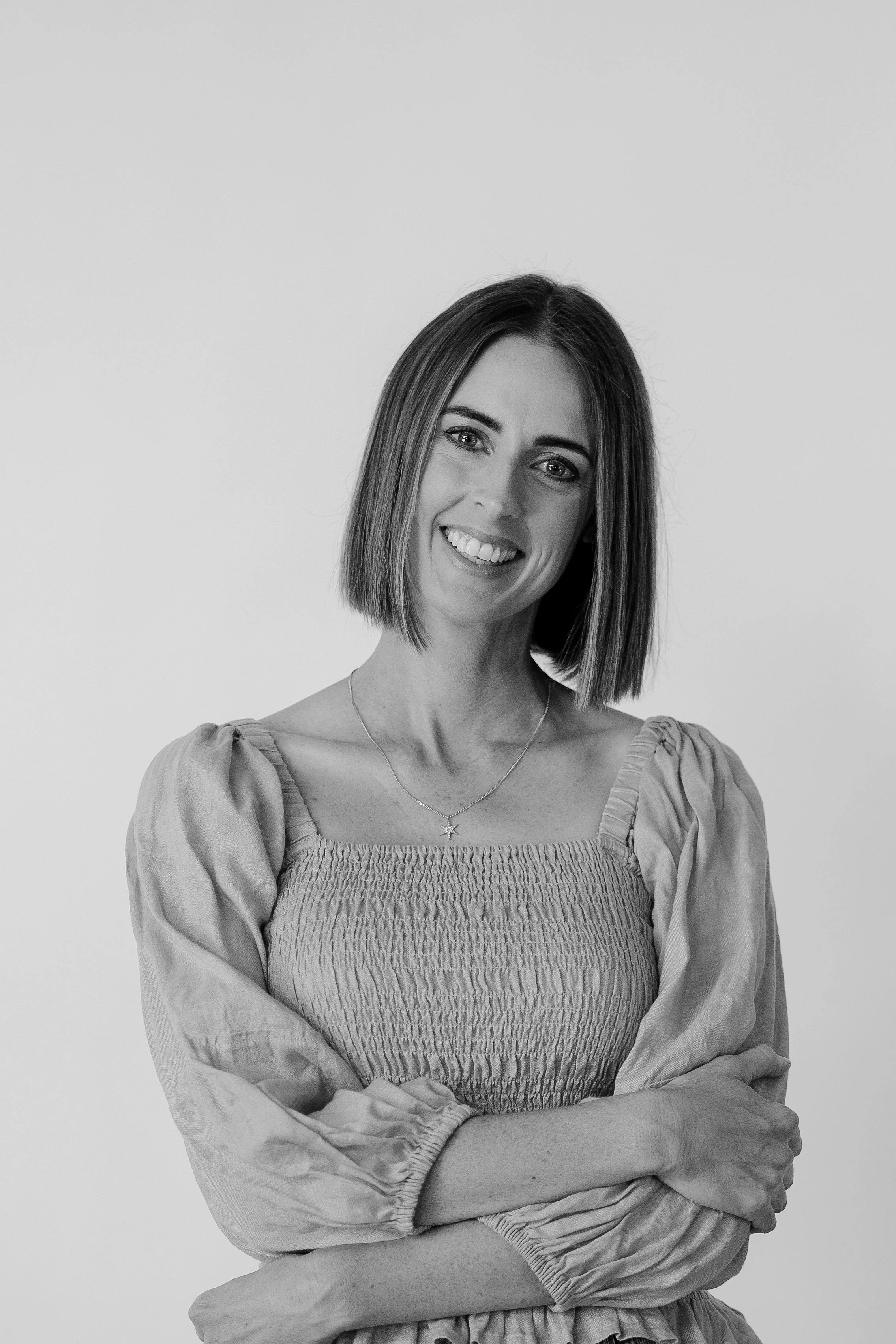 A woman with shoulder-length hair smiling, wearing a textured dress with puffed sleeves, crossed arms, and a star-shaped necklace against a plain background.