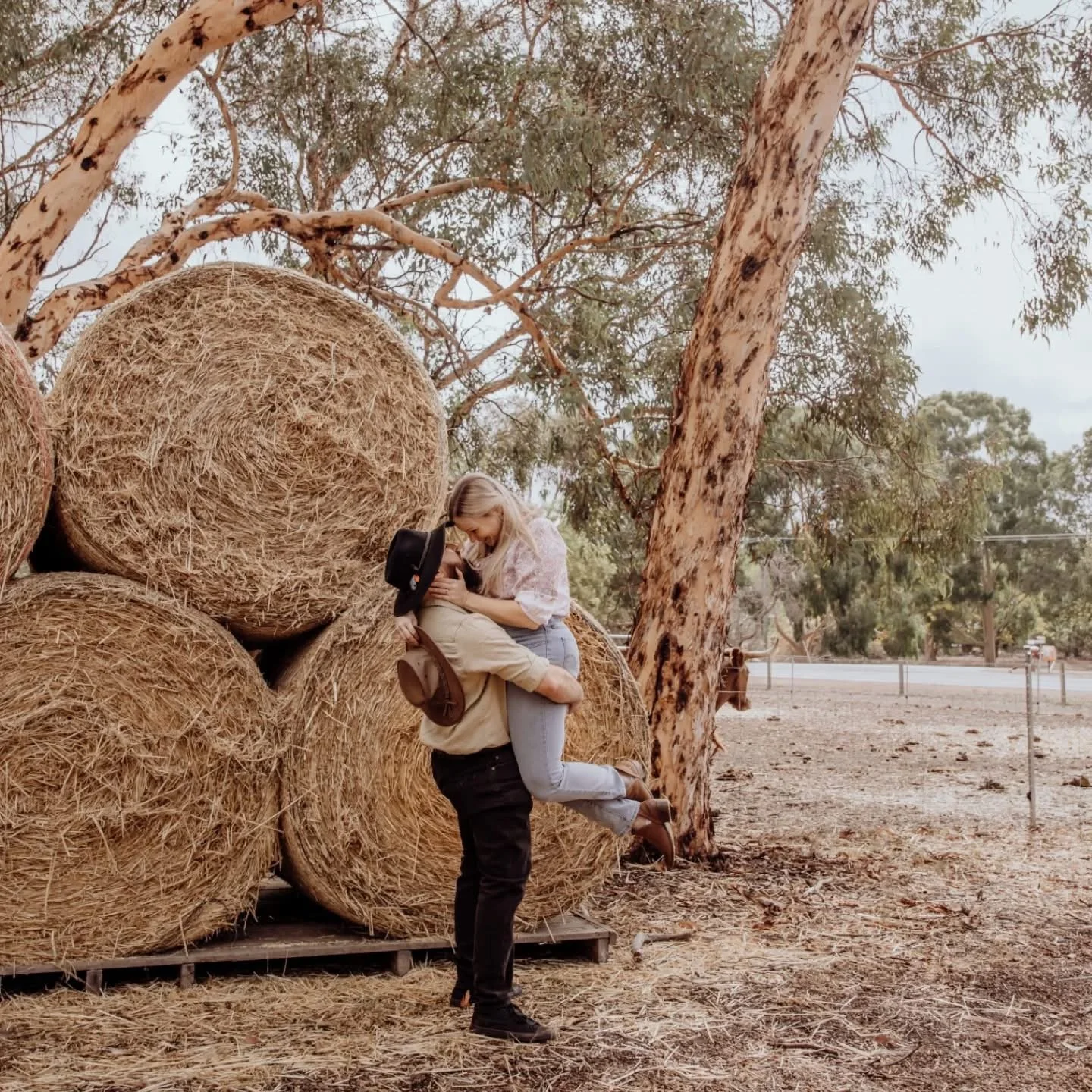 A couple is near a stack of large hay bales in an outdoor rural setting, with a woman in a light top and jeans sitting on the man, who is standing. They are embracing affectionately, with one holding a black hat and another holding a brown hat. There are trees and a road with a fence in the background.