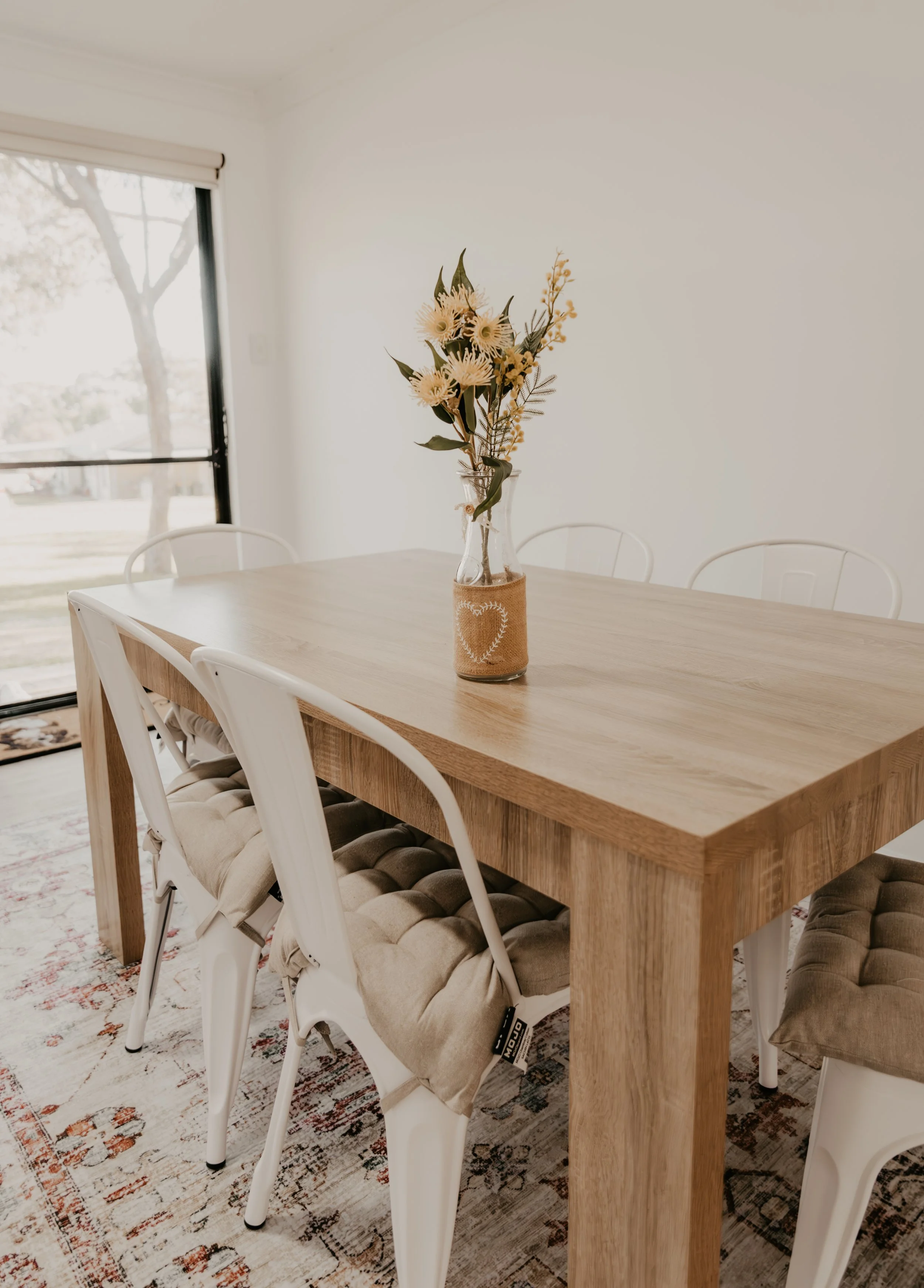 A wooden dining table with six white chairs around it, a vase with beige and green artificial flowers in the center, on a rug near a sliding glass door with a tree outside.