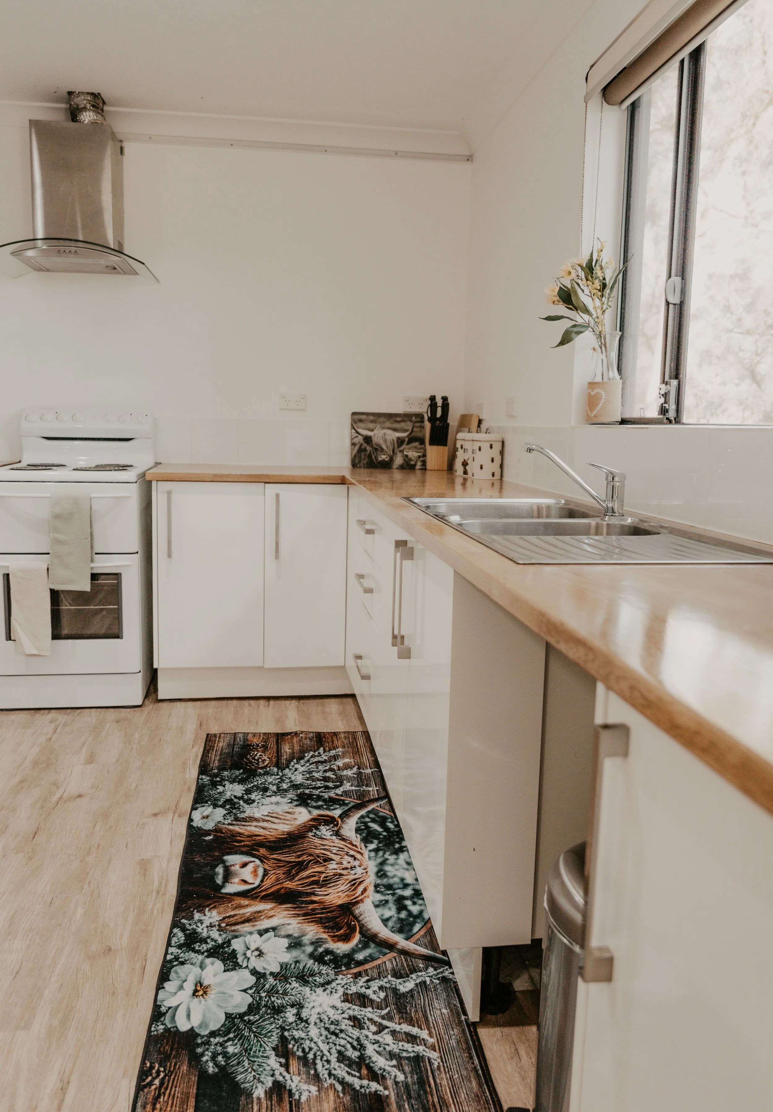 A kitchen with white cabinets, a wooden countertop, a stainless steel sink, a white stove, a decorative tiger rug on the wooden floor, and a bouquet of flowers in a vase near a window.
