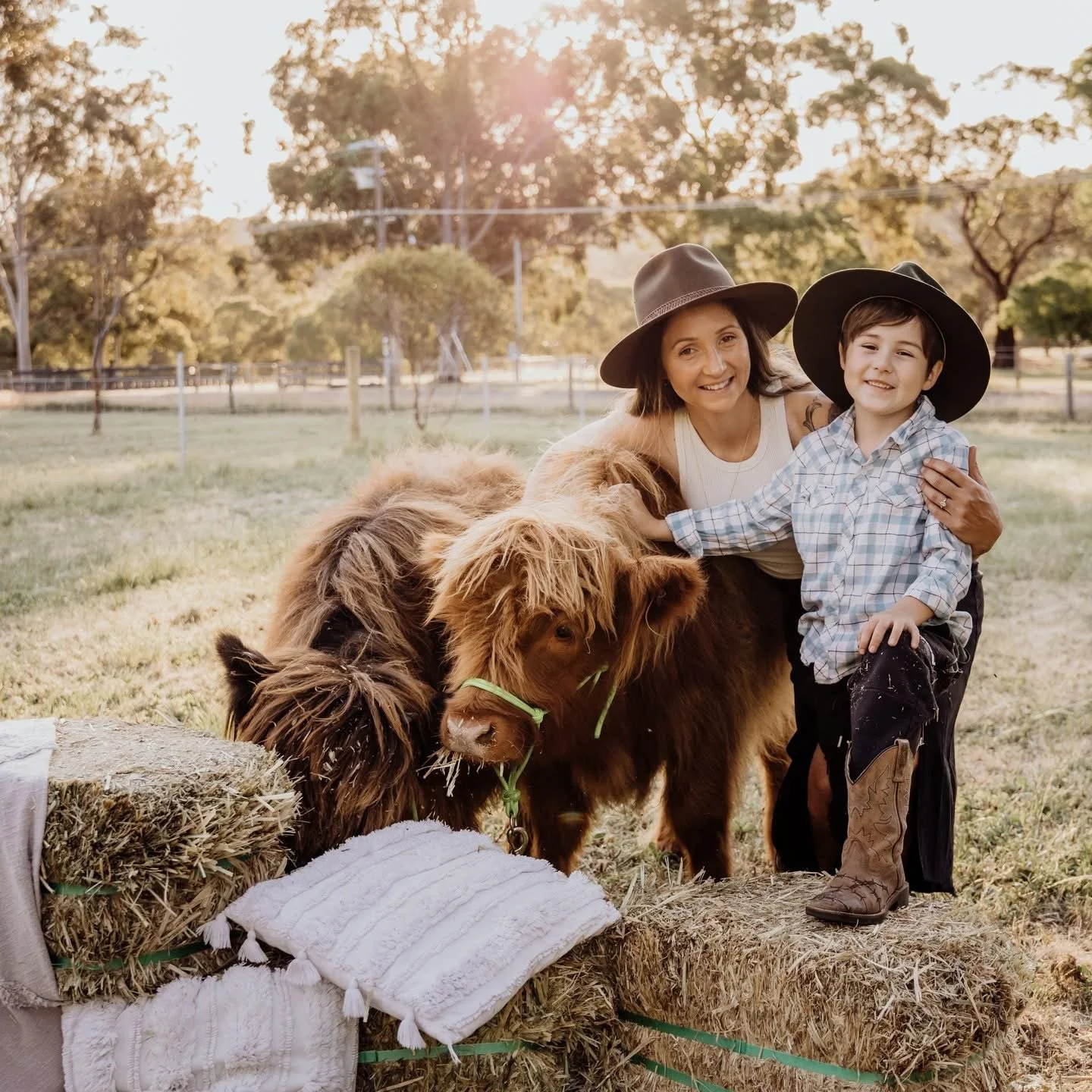 A woman and a young boy smiling and hugging a brown Highland cow in a grassy outdoor park during sunset. The woman is wearing a hat and a sleeveless top, while the boy is dressed in a plaid shirt and cowboy boots, both wearing wide-brimmed hats.