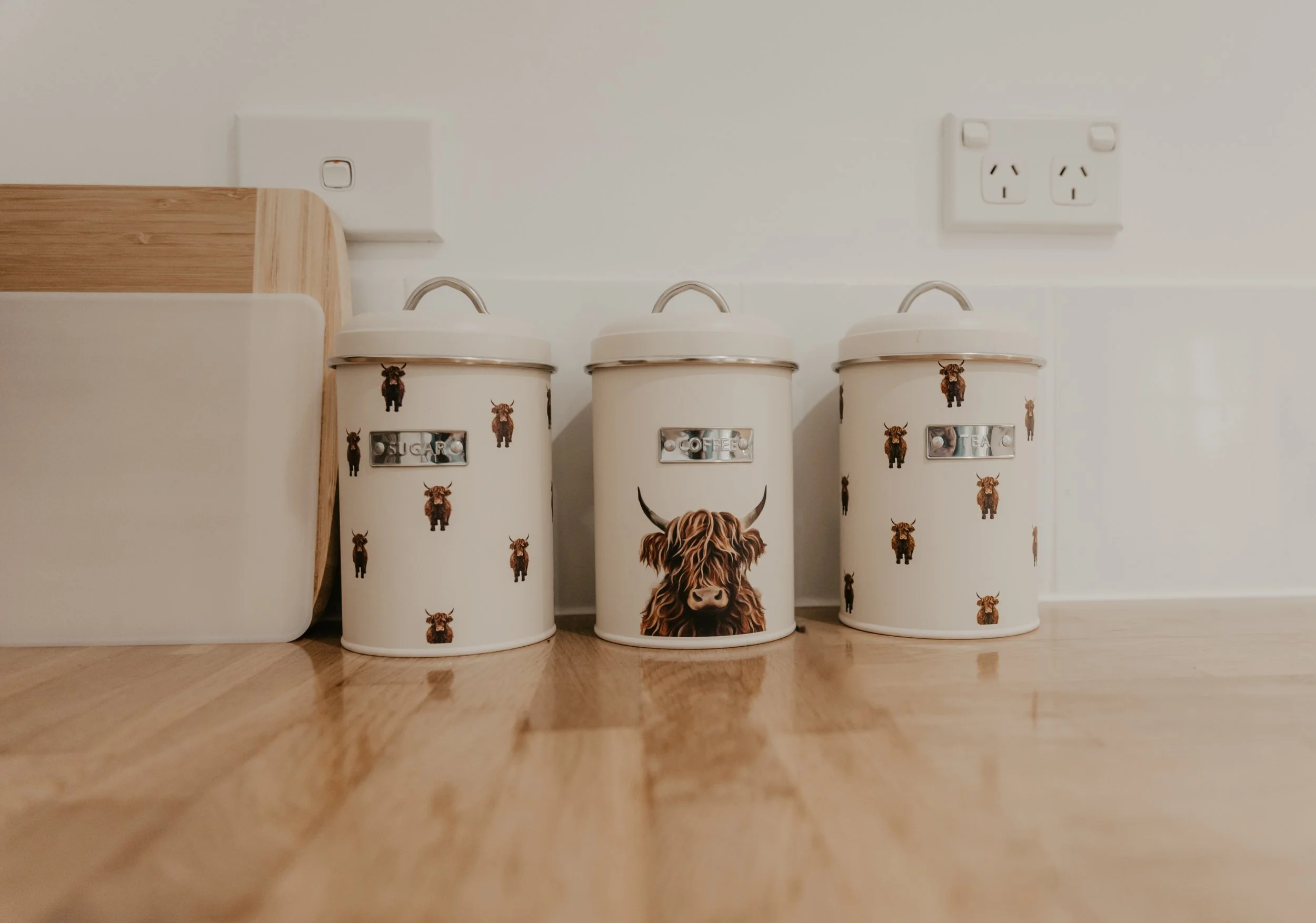 Three white kitchen canisters with black cow patterns and labels for sugar, coffee, and tea, placed on a wooden floor against a white wall with electrical outlets.