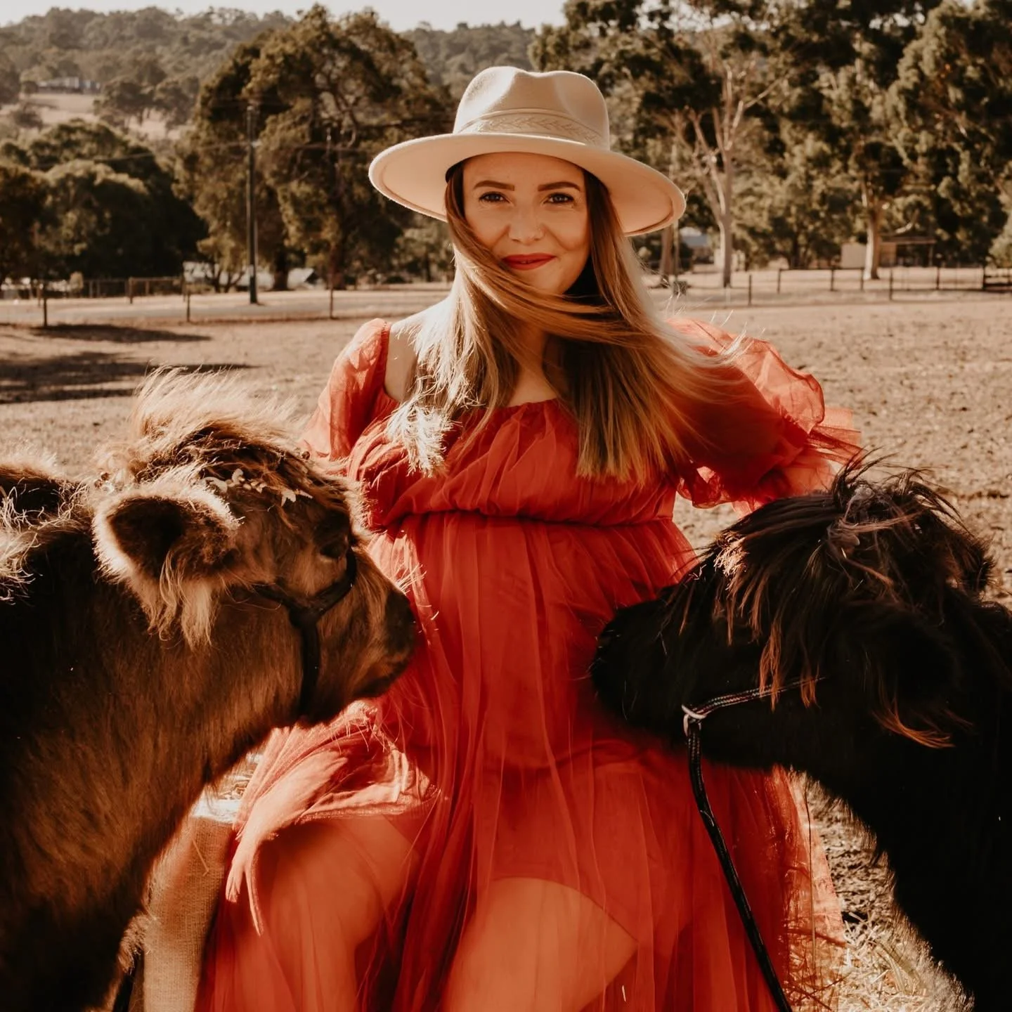 A woman with long hair wearing a red dress and a white wide-brimmed hat standing outdoors among trees, with two small horses near her, one on each side.