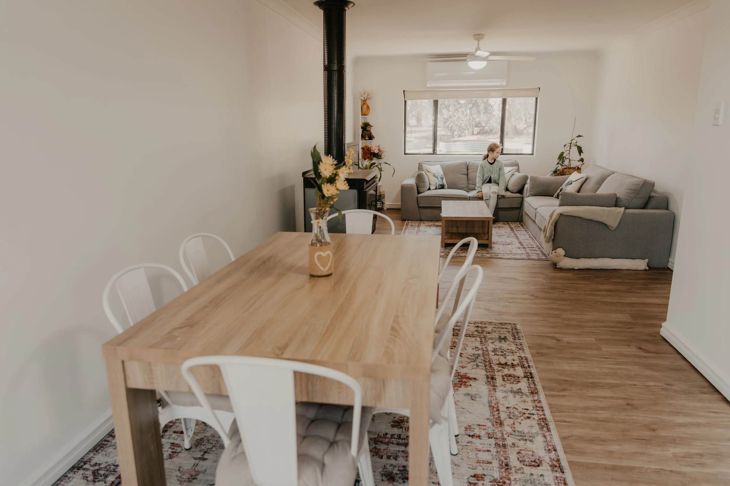 A dining area with a wooden table and white chairs in the foreground, and a living room with a gray sofa, a coffee table, and decorative plants near the window in the background. The room has hardwood floors and a patterned rug under the coffee table.