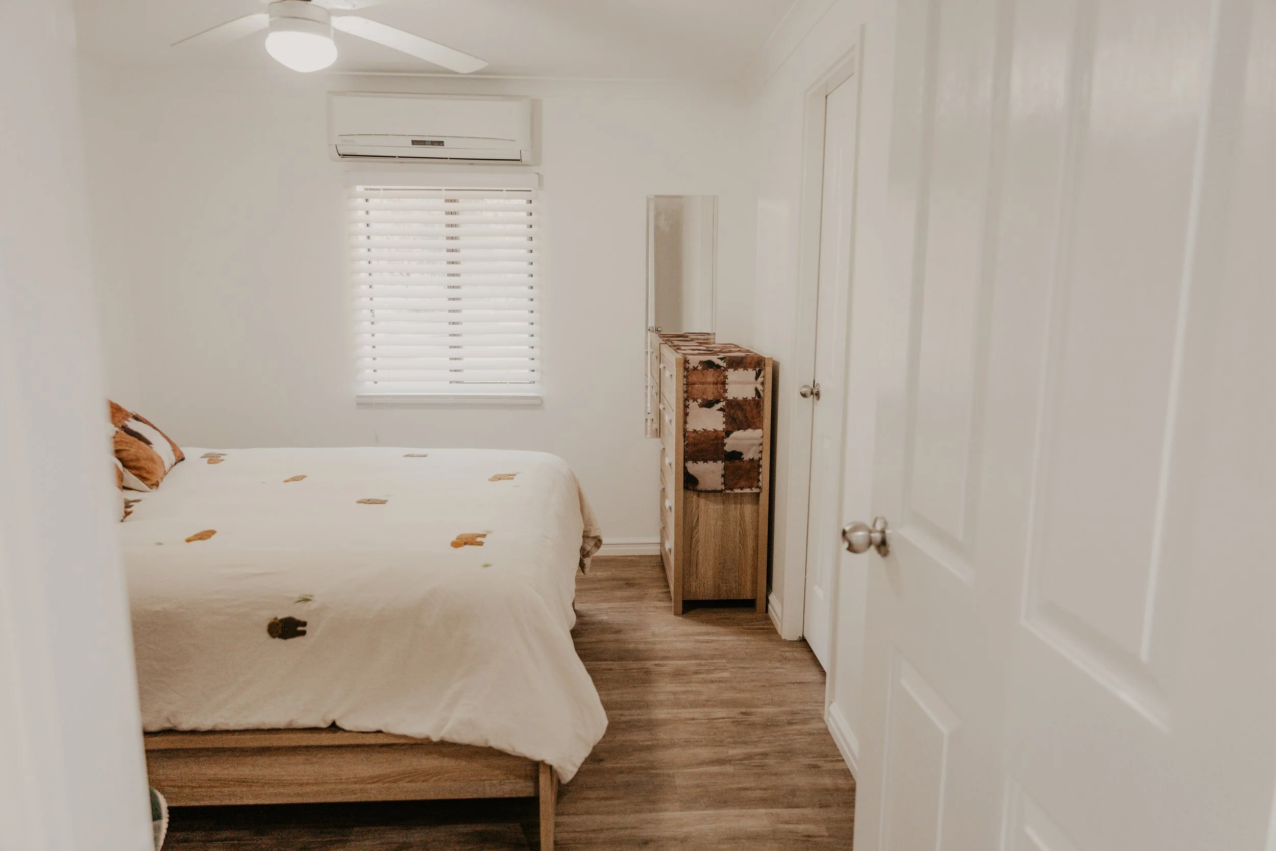 A bedroom seen through an open white door. The room has a bed with a white comforter that has small animal prints, a window with white blinds, a ceiling fan, a wooden dresser with a mirror, and white walls with a split air conditioning unit above the window.