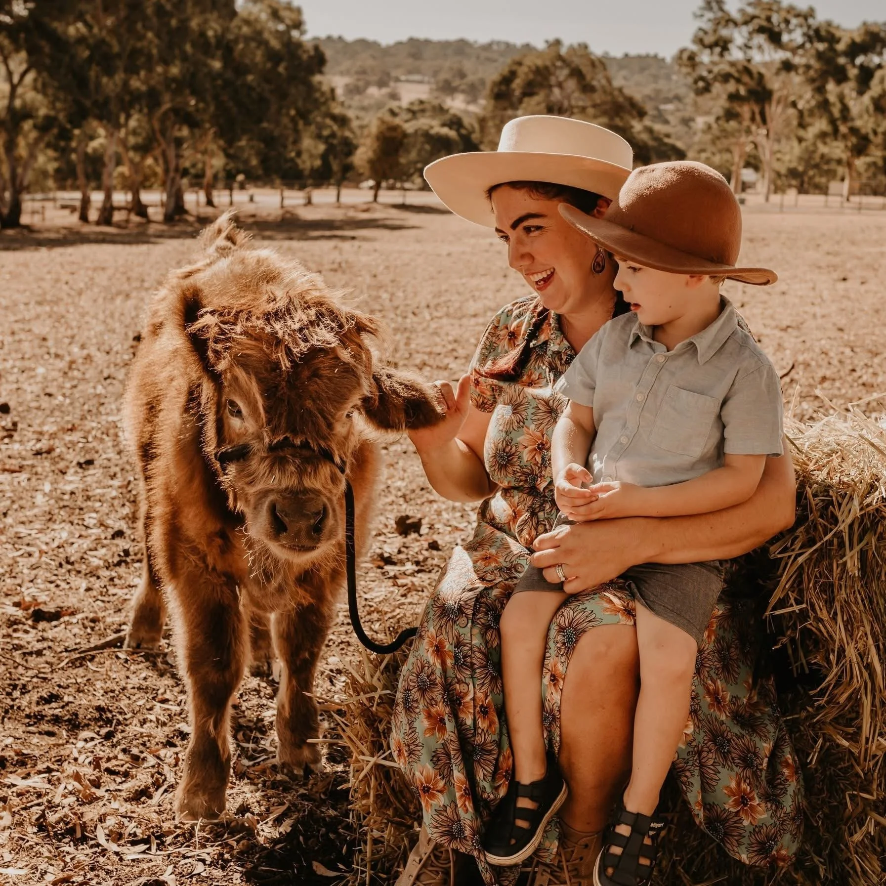 Woman and young boy with sun hats smiling and petting a baby lion in a park or farm setting.