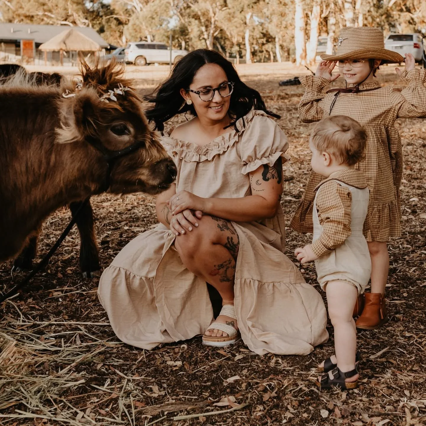 A woman with dark hair and glasses with freckles, sitting on the ground in a beige dress, smiling at a brown cow with flowers in its hair, surrounded by two young children with blonde hair and wearing vintage-style plaid dresses outdoors with trees and parked cars in the background.
