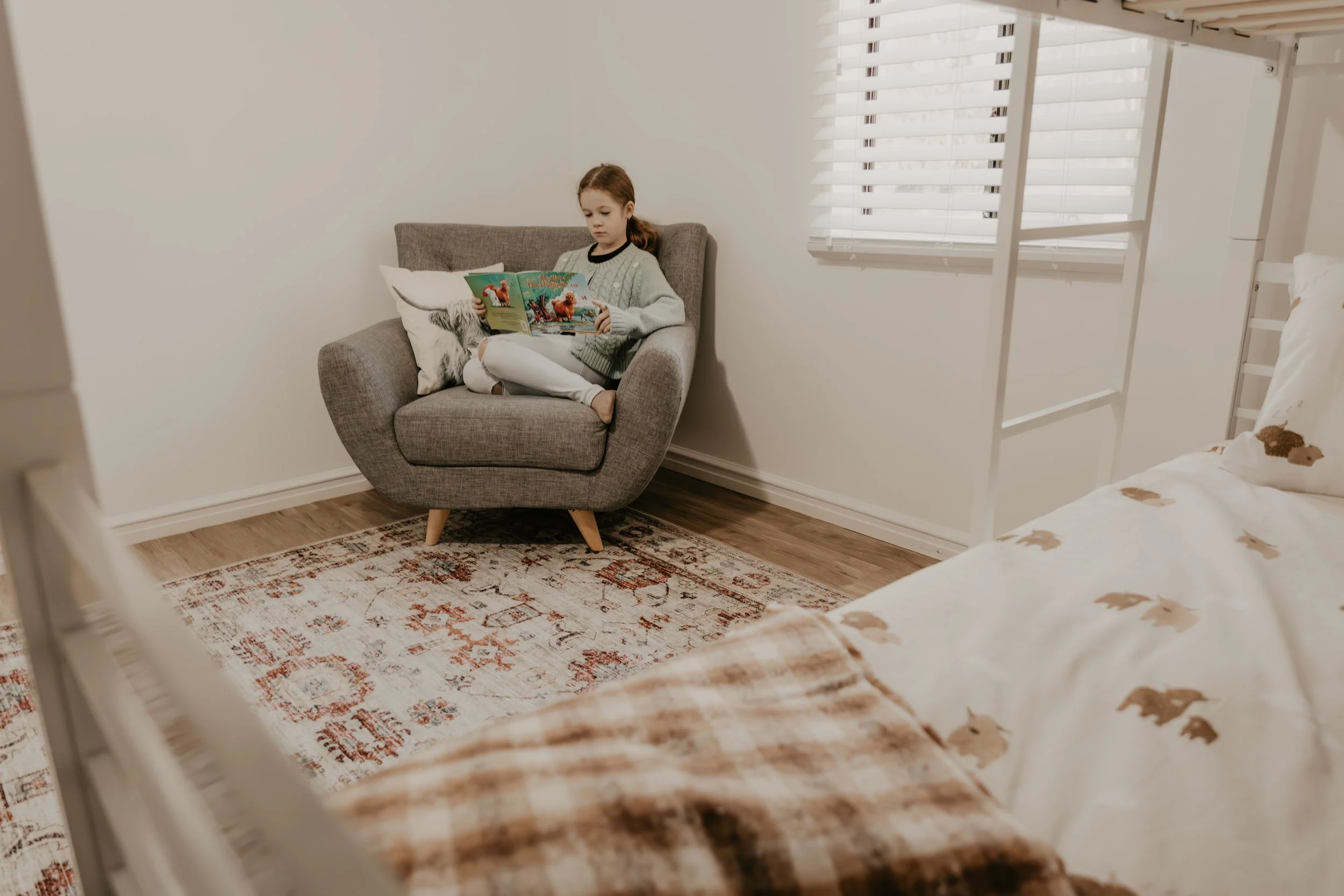 A young girl with brown hair tied back, sitting cross-legged on a gray armchair, reading a children's book titled 'The Ugly Duckling' in a bright room with white walls and blinds, a bed in the foreground, and a patterned rug on the wooden floor.