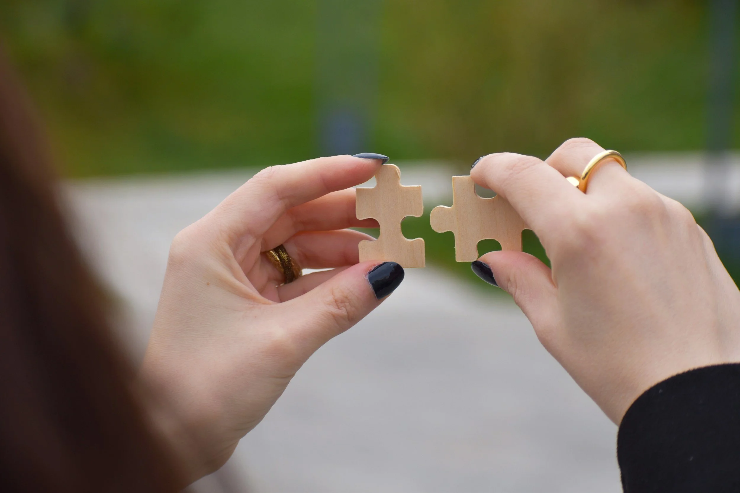 Two hands with dark nail polish holding a wooden puzzle piece, connecting with another piece, with blurred green background.