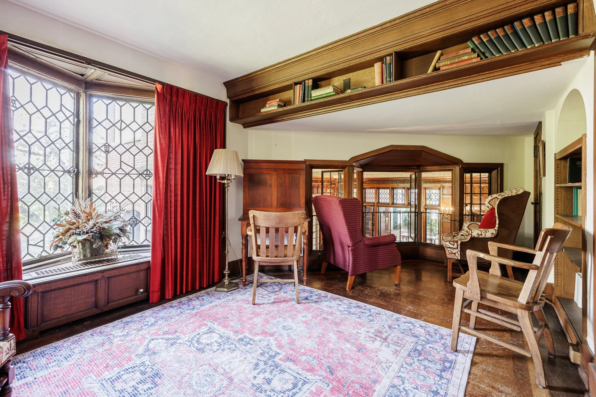 Living room with wooden furniture, red curtains, patterned rug, and multiple chairs and shelves.