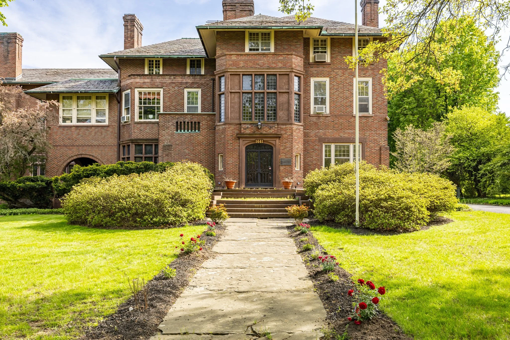 A large brick mansion with multiple stories and a front porch, surrounded by green trees and a well-maintained lawn. A stone pathway leads to the front door, which is flanked by two potted plants.