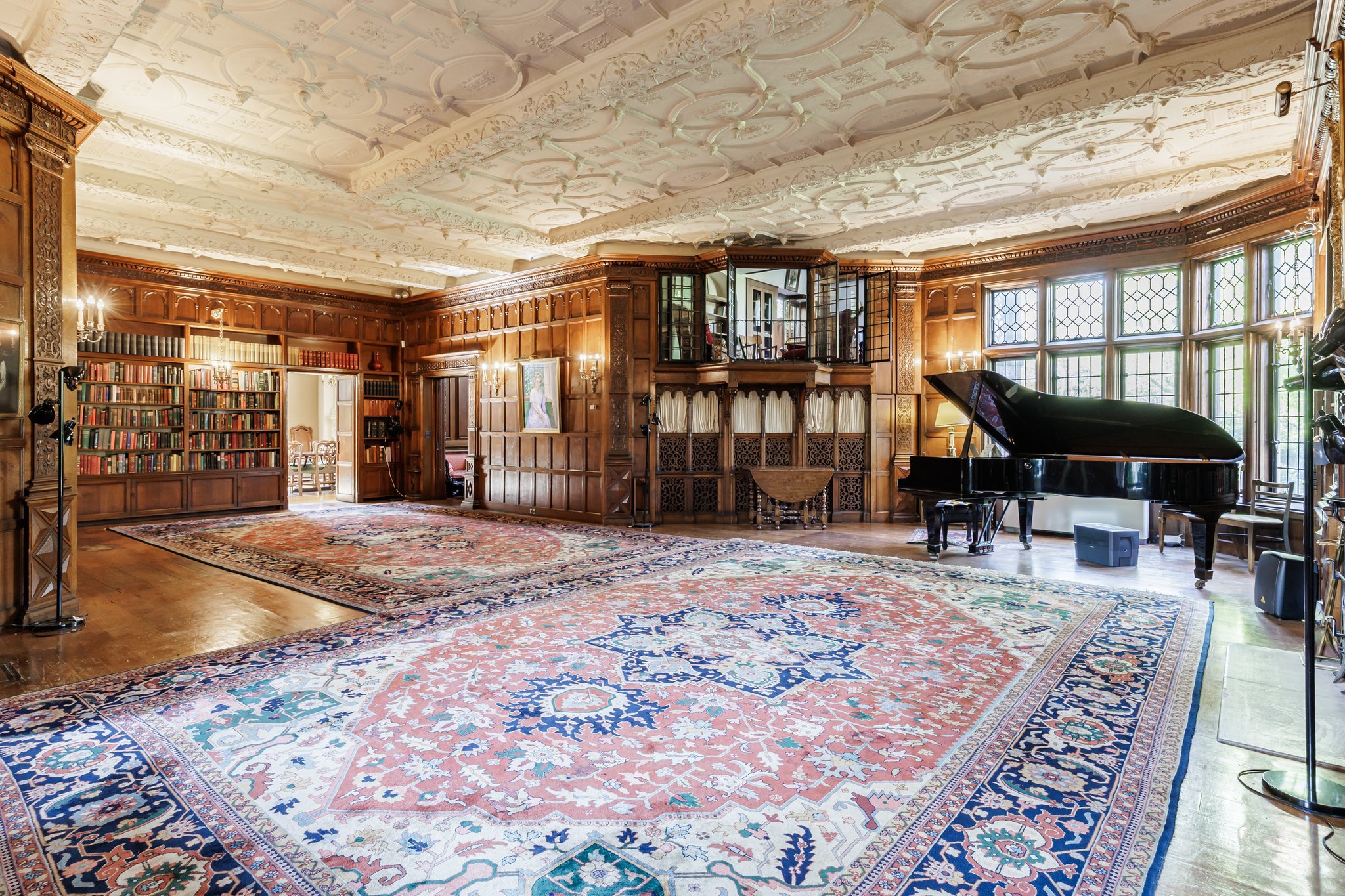 Elegant living room with ornate ceiling, wooden paneling, large windows, a grand piano, shelves filled with books, a portrait painting, chandeliers, and a large patterned area rug.