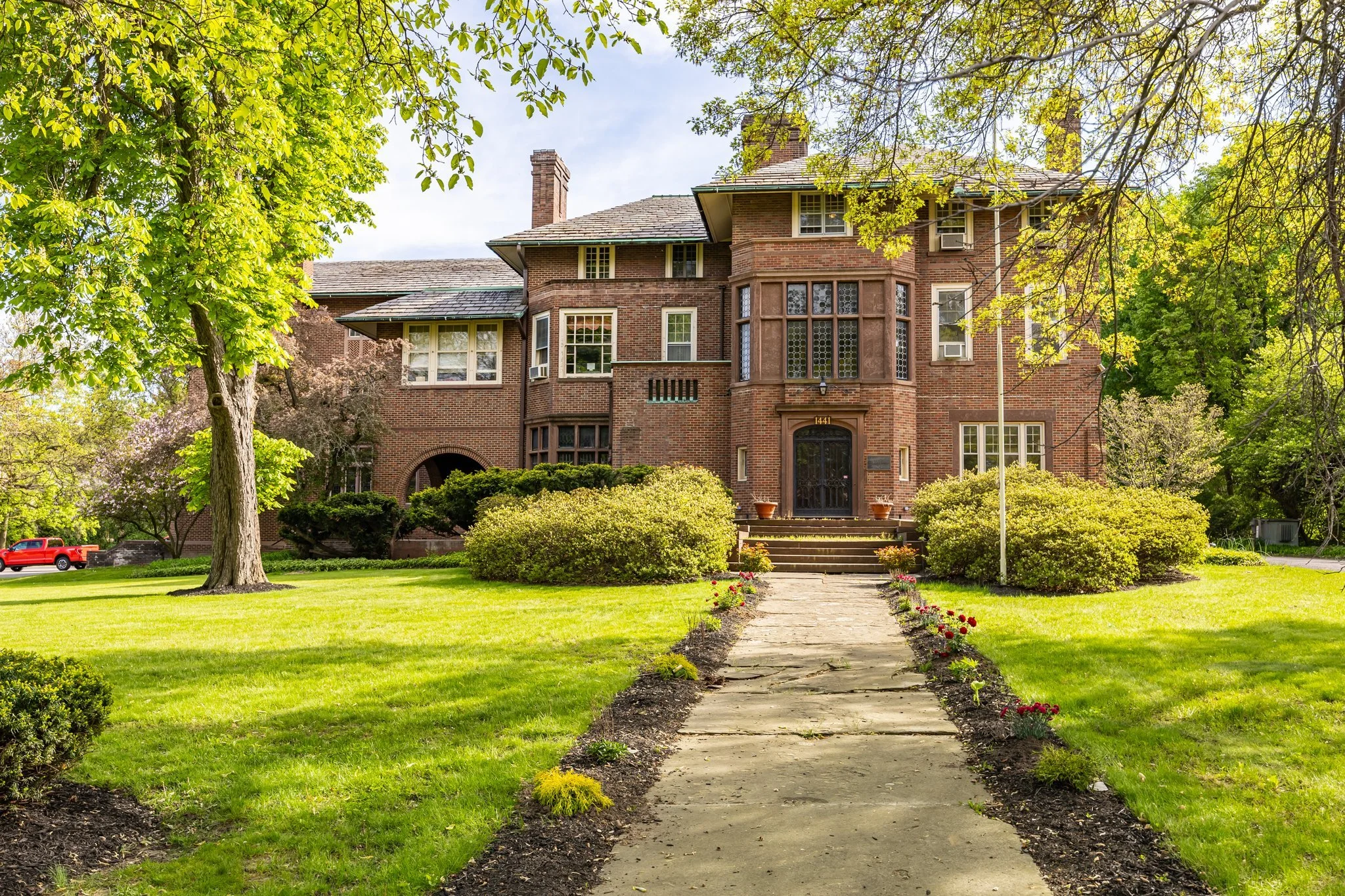 A large brick mansion with multiple stories, bay windows, and a front porch, surrounded by a well-maintained garden with a stone pathway, green lawn, trees, and shrubs, under a clear blue sky.