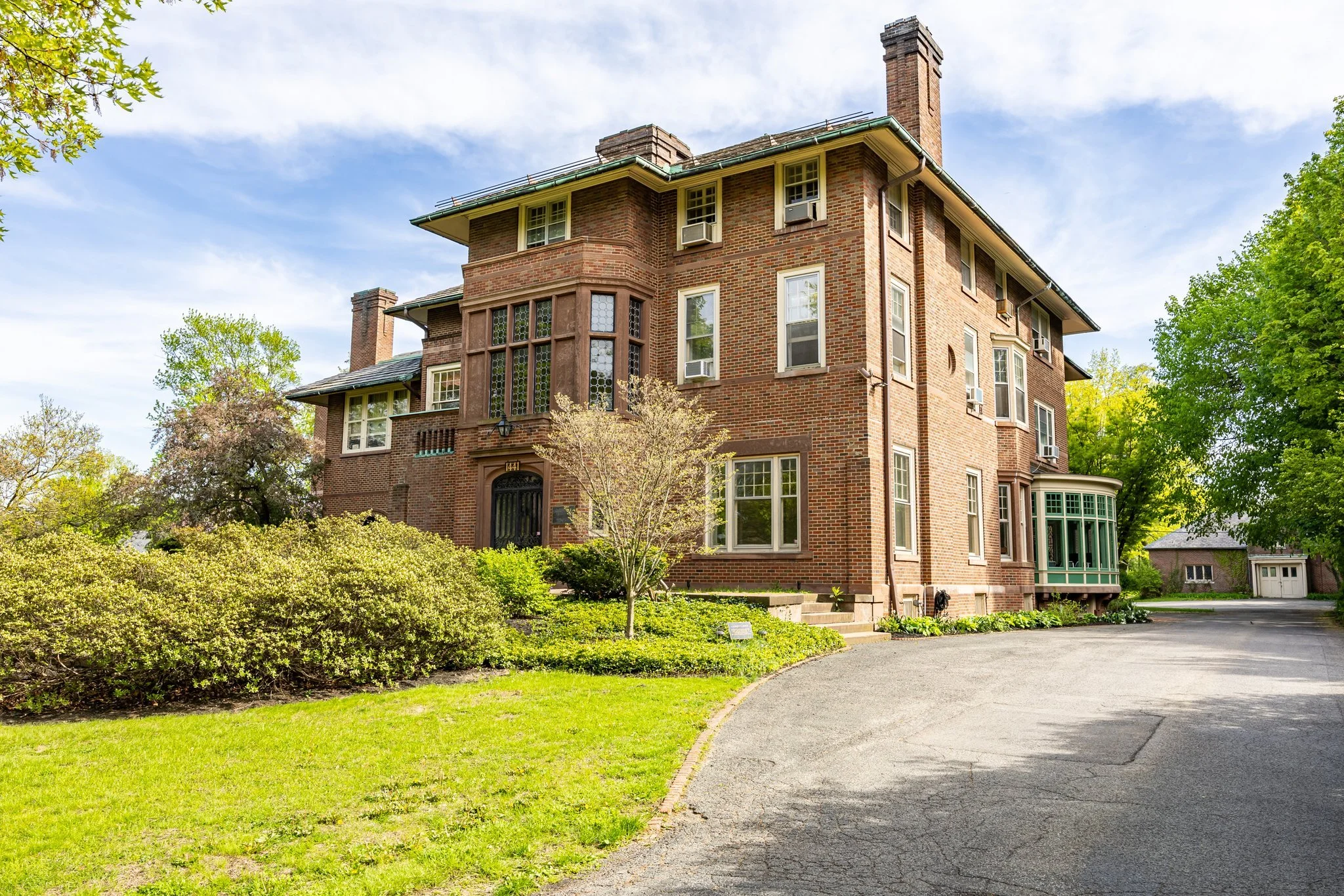 A large mansion with multiple windows, air conditioning units, and a tall chimney. The mansion is surrounded by green trees and shrubs, with a curved driveway leading up to the entrance. The sky is partly cloudy.