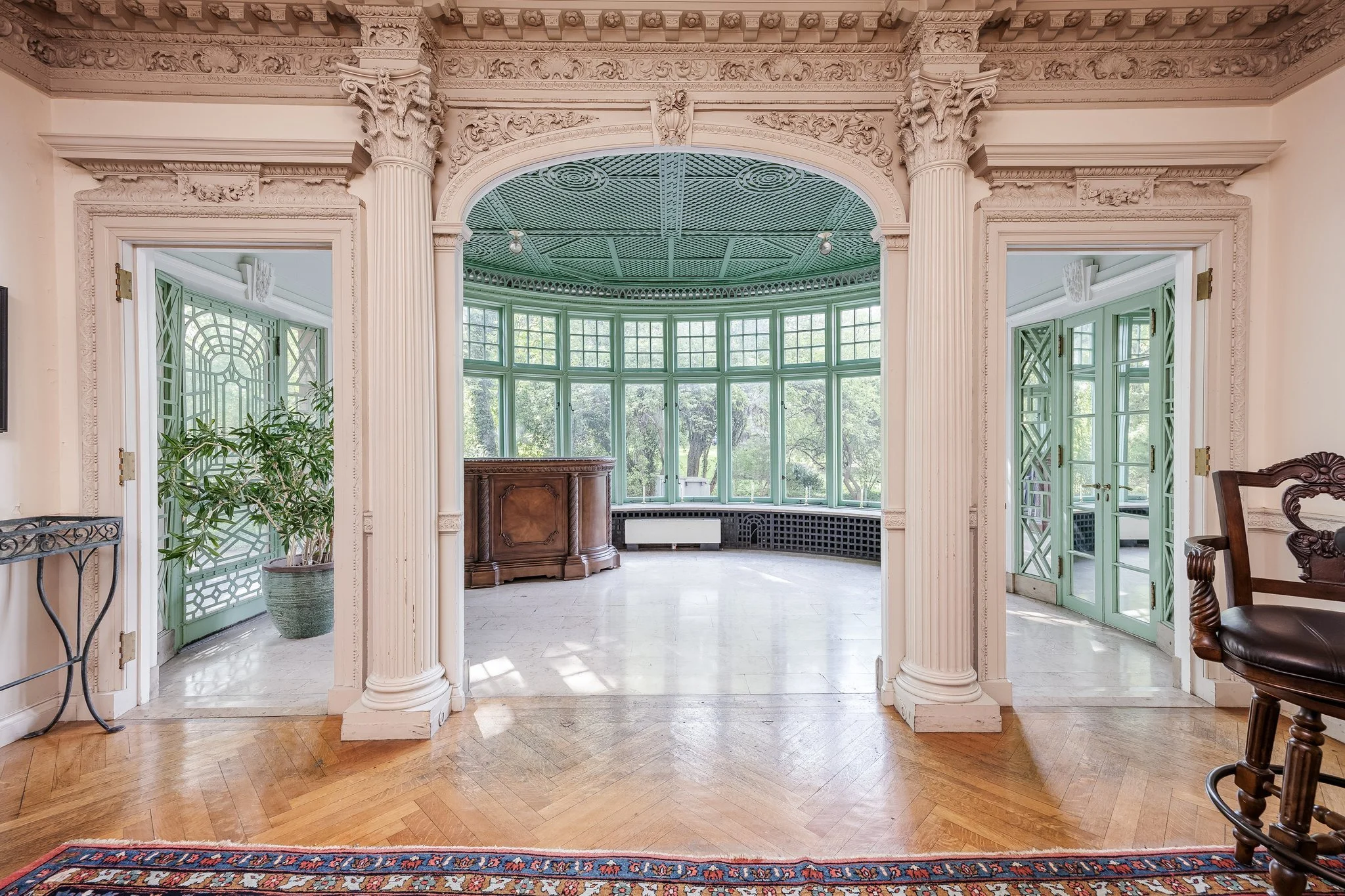 An ornate stone archway with decorative classical columns leading to a room with large green window frames and a curved glass wall, featuring a patterned ceiling and natural light.