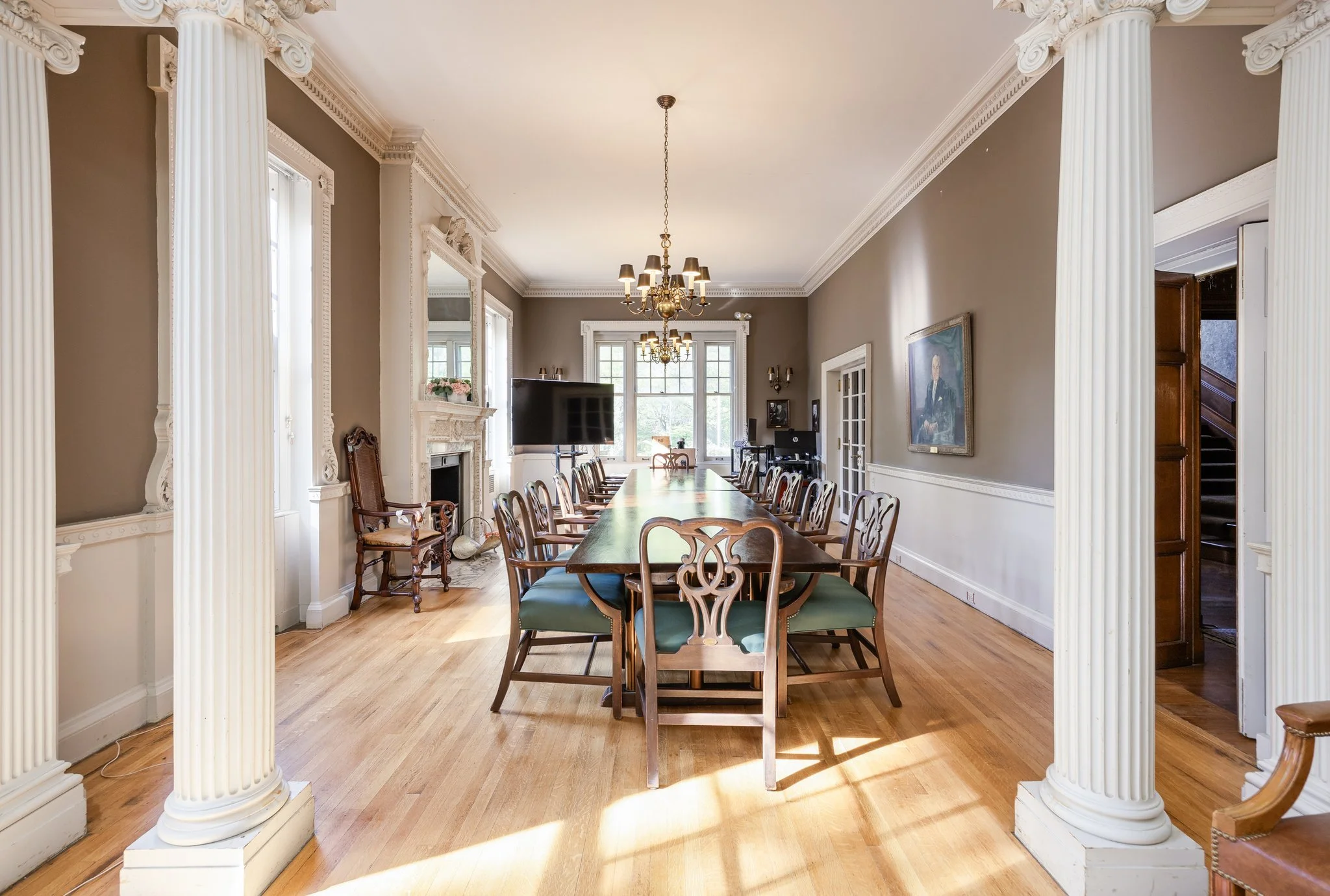 Interior of a dining room with a long wooden table and chairs, ornate white columns, and large windows with curtains providing natural light. There are chandeliers hanging from the ceiling, and a portrait hanging on the wall. The room has beige walls and hardwood floors.